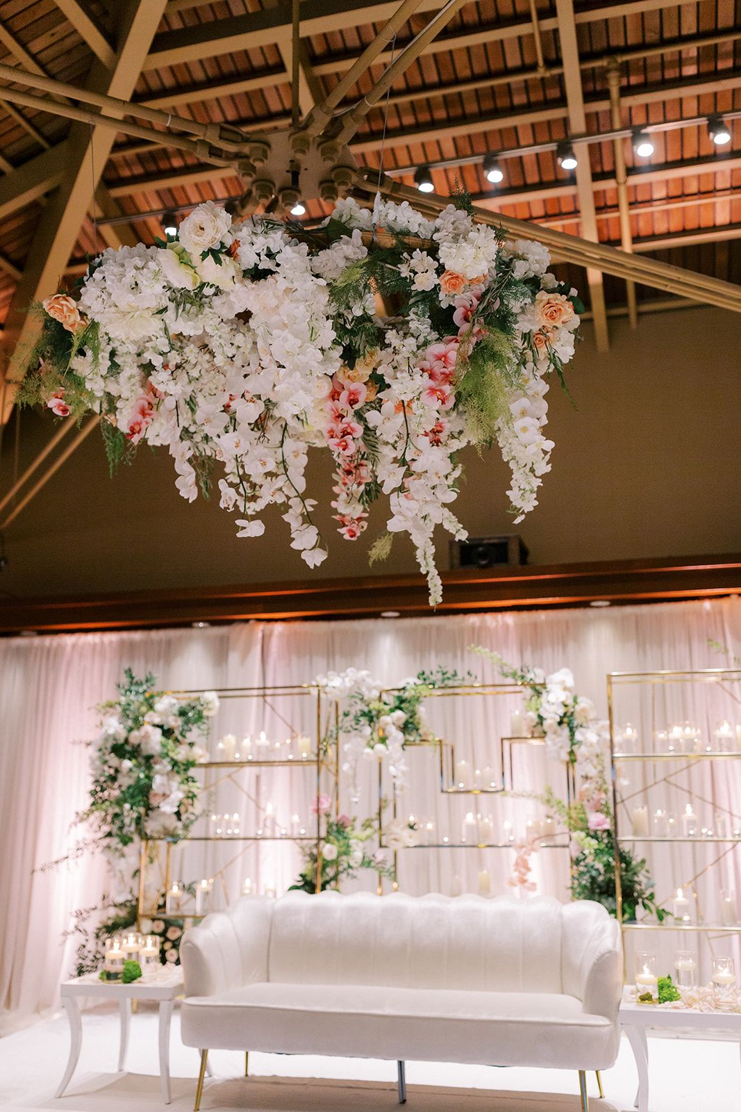 Elegant wedding reception stage featuring a white lounge sofa, suspended floral installation, and romantic floral backdrop.