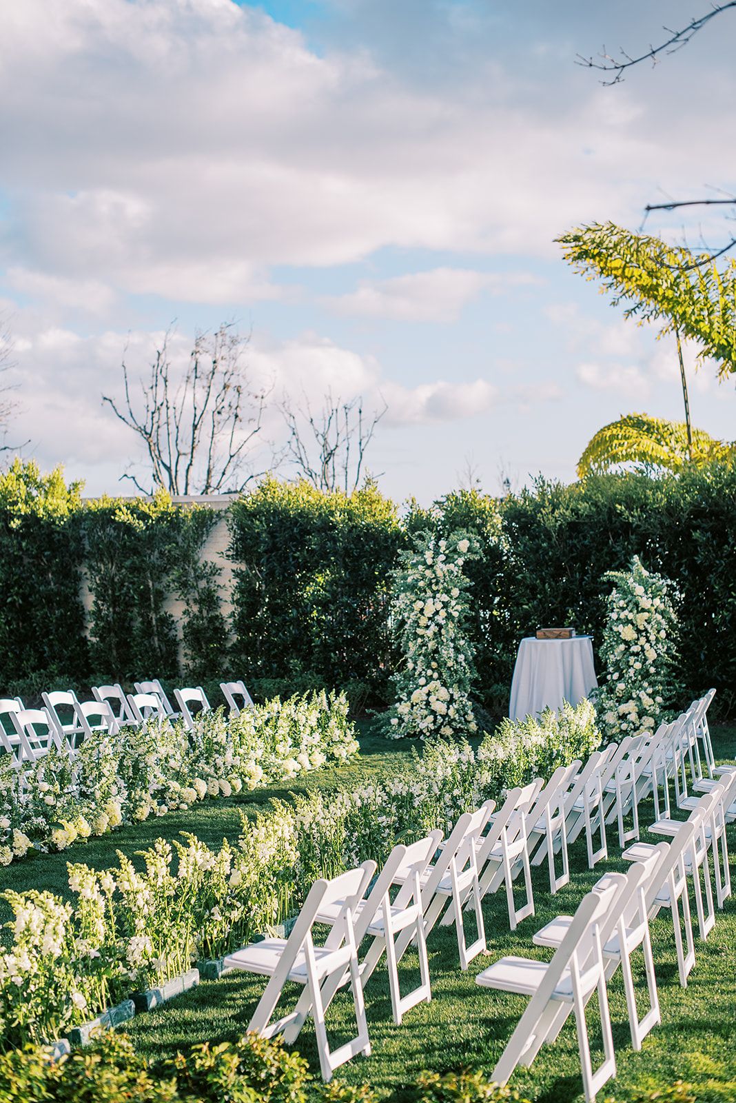 Outdoor wedding ceremony setup with white chairs, lush greenery, and elegant white floral aisle decor in a garden venue