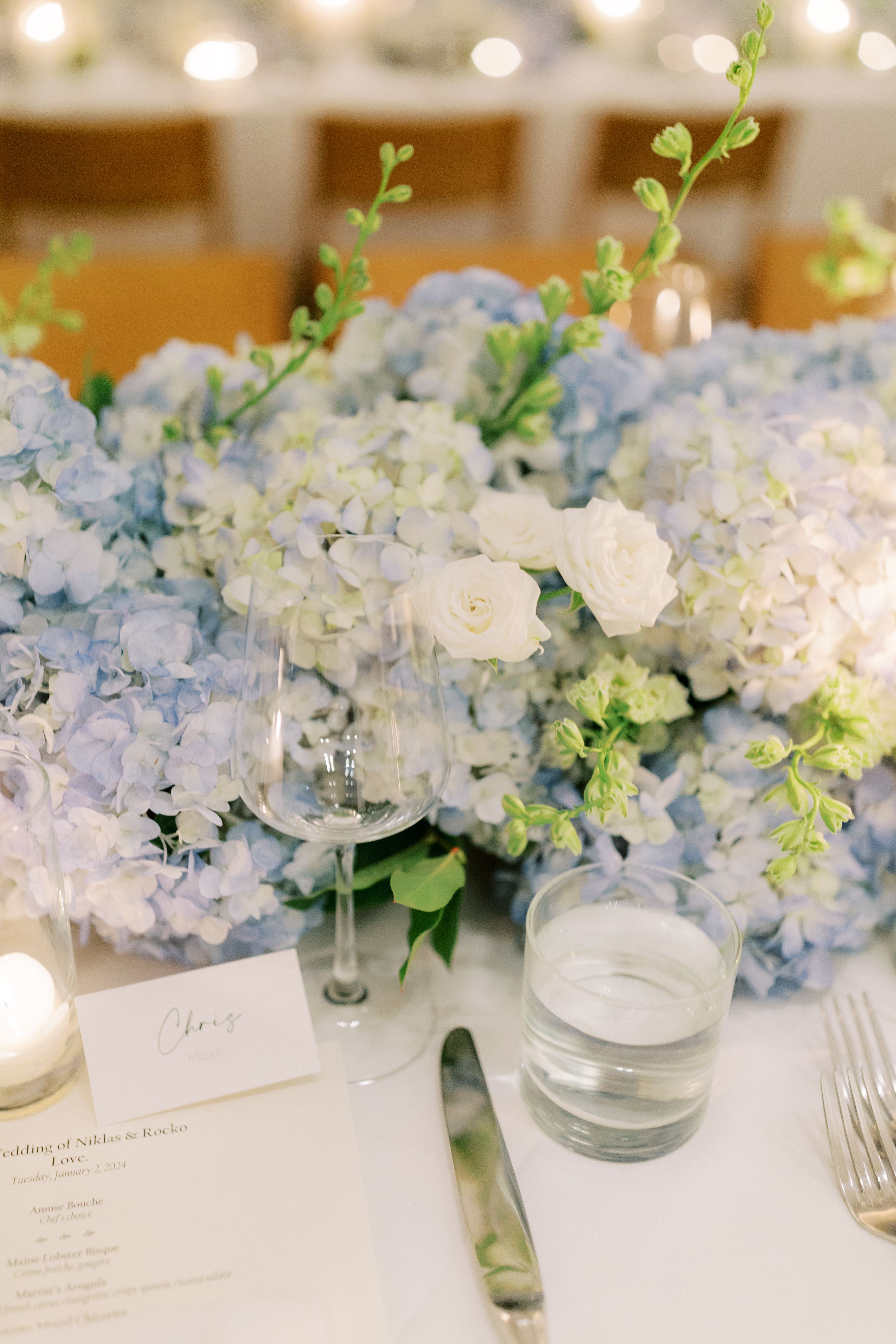 Luxury wedding reception table featuring blue hydrangea floral arrangements, white roses, candles, and elegant place settings.