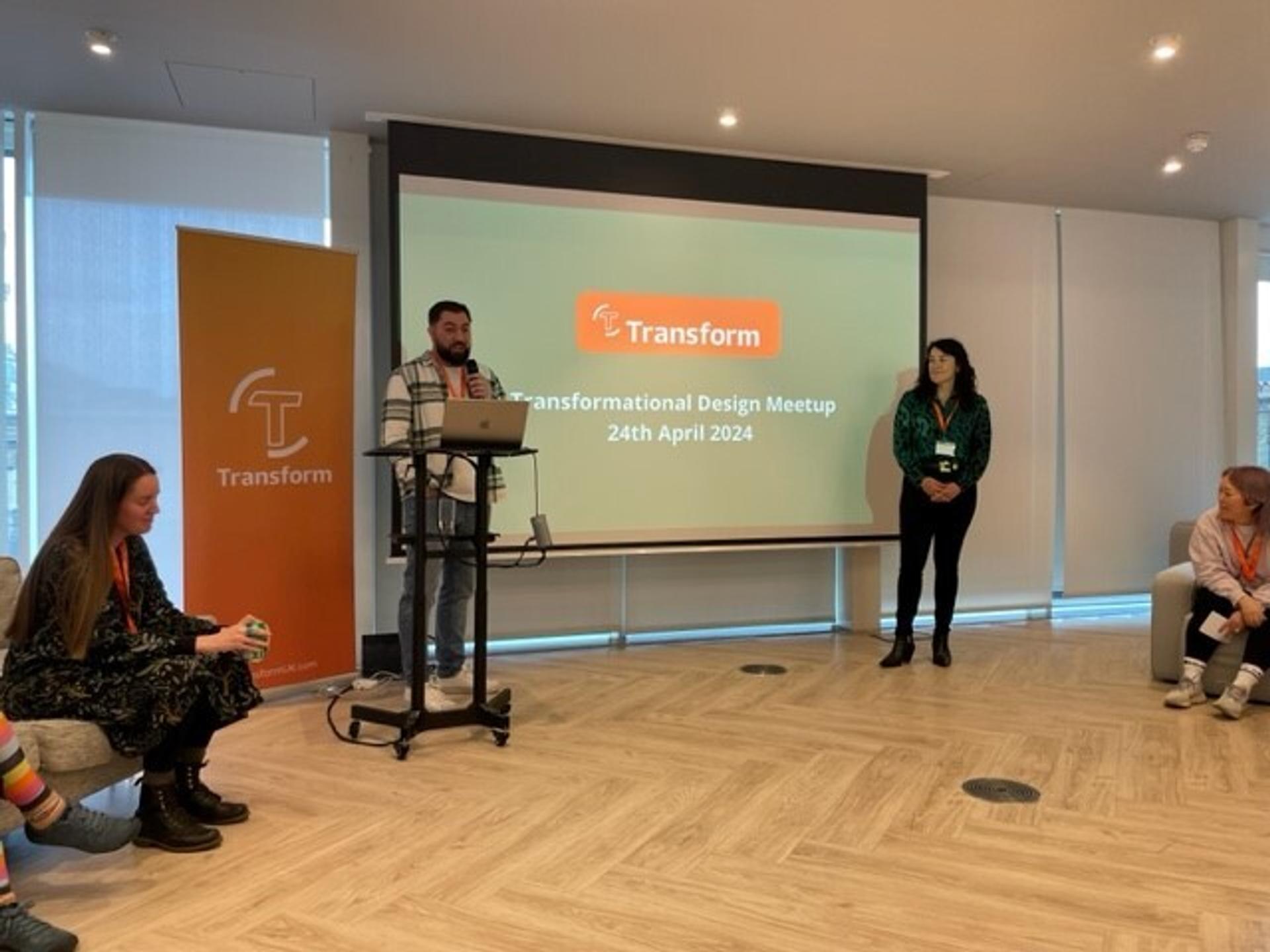 A man standing behind a podium with a woman a little ways off - both are in front of a screen that reads 'Transformational Design Meetup 24th April 2024'