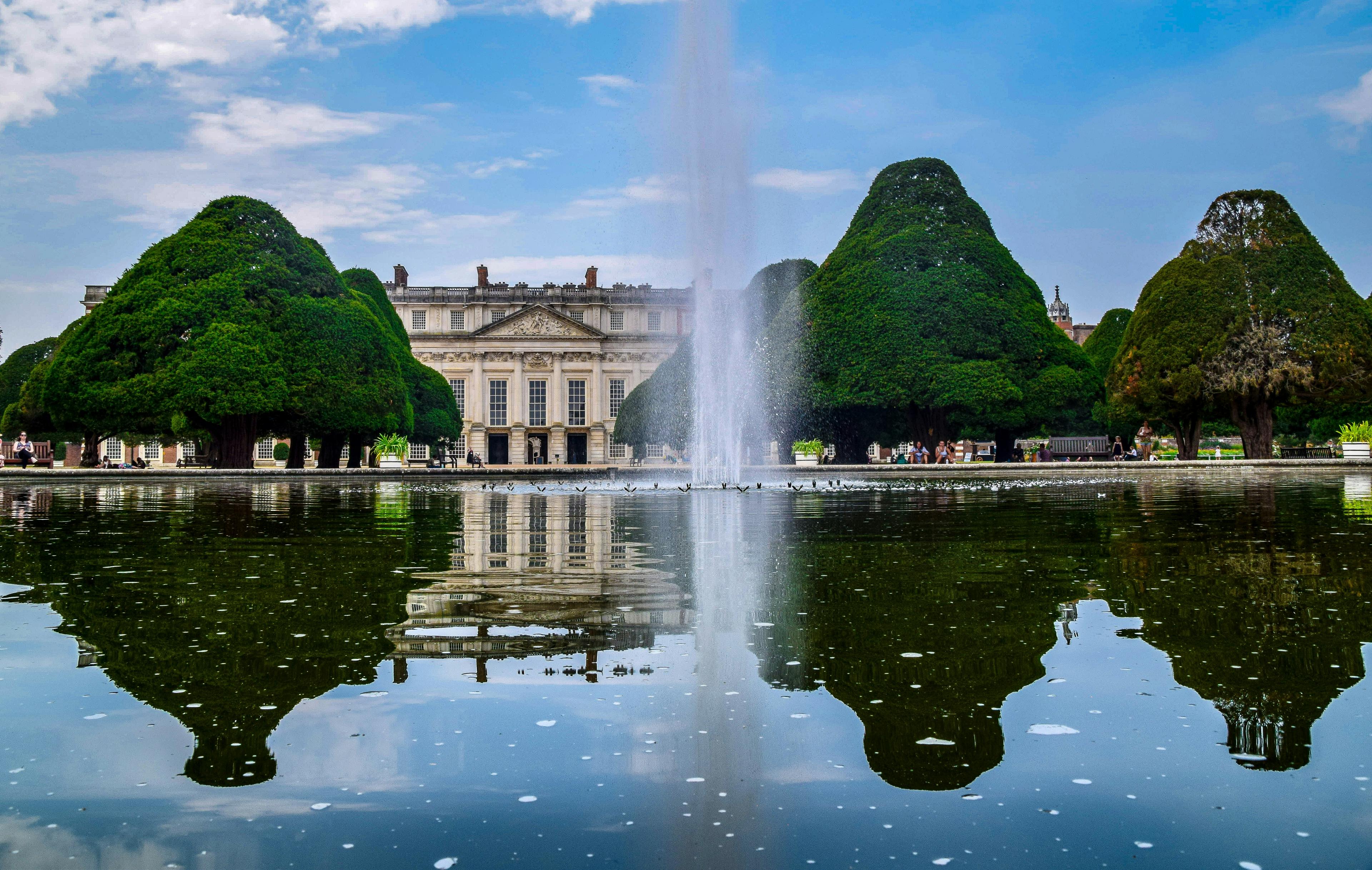 A spurting fountain on a lake with trees and Kensington Palace in the background