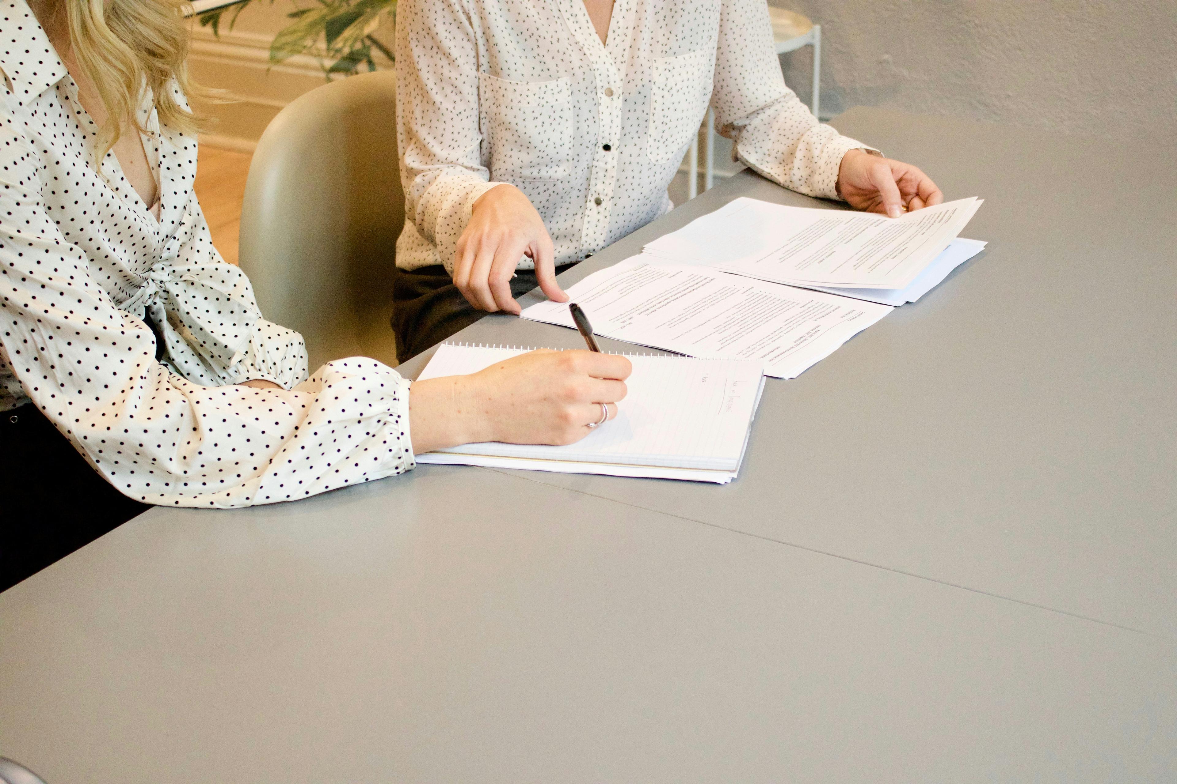 Two women seated at a table. One of them is paging through a stack of papers and another is taking notes.