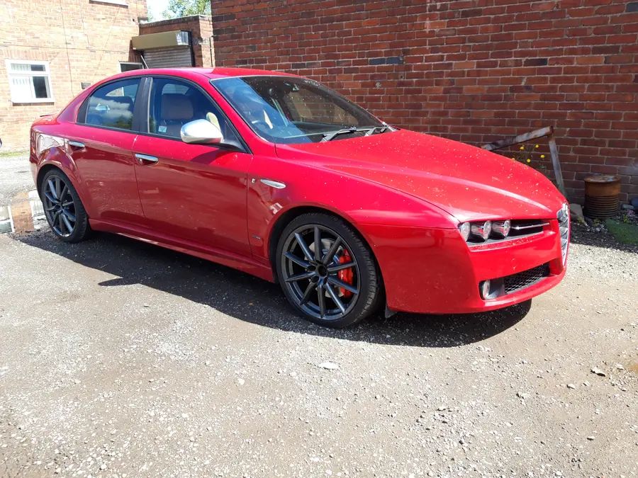 A red Alfa Romeo 159 on gloss black wheels with red calipers, against a brick wall