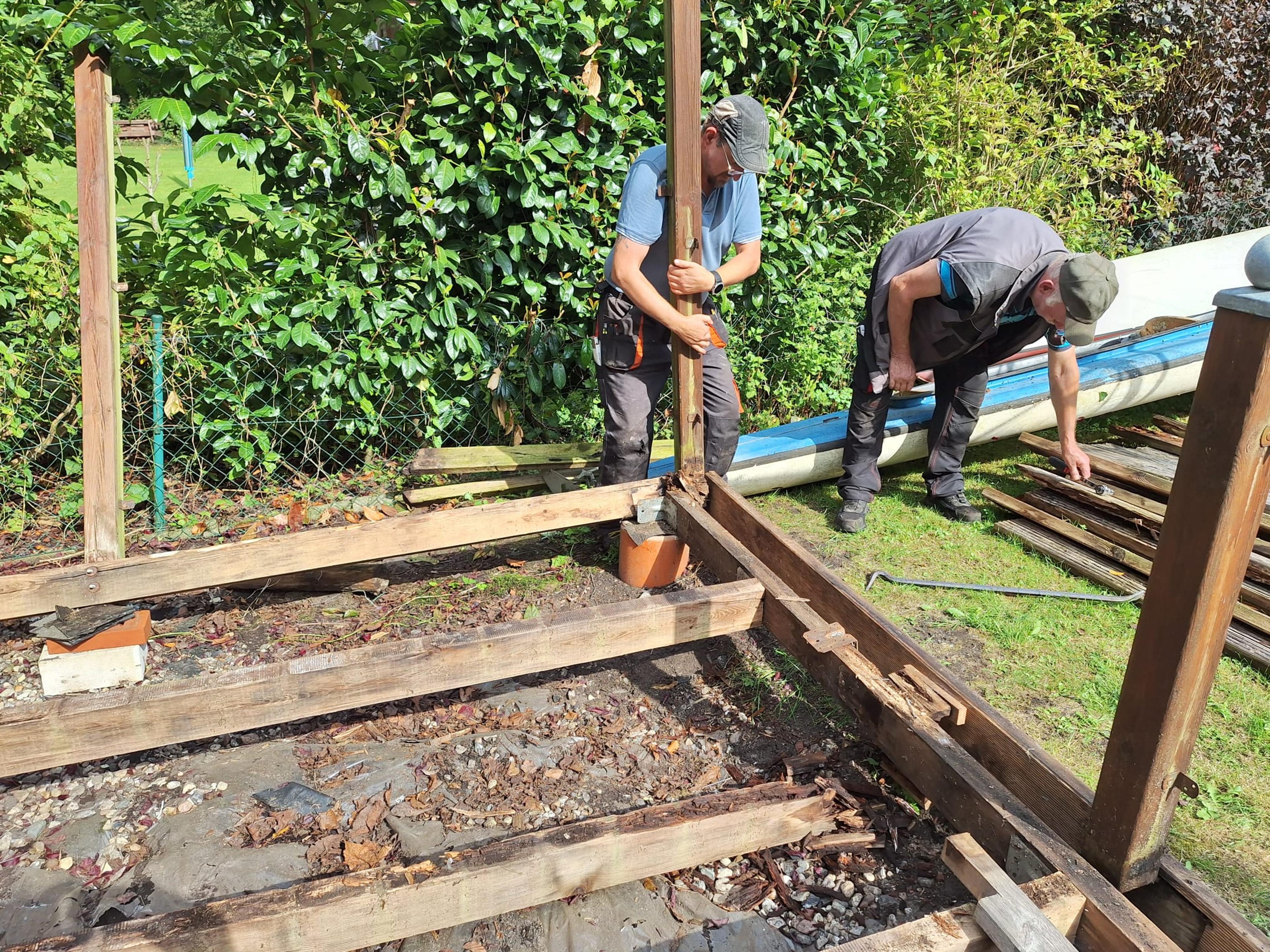 Zwei Männer bauen im Garten einen Holzzaun, einer hält einen Pfosten, der andere sortiert Holzplanken. Im Hintergrund sind grüne Büsche.