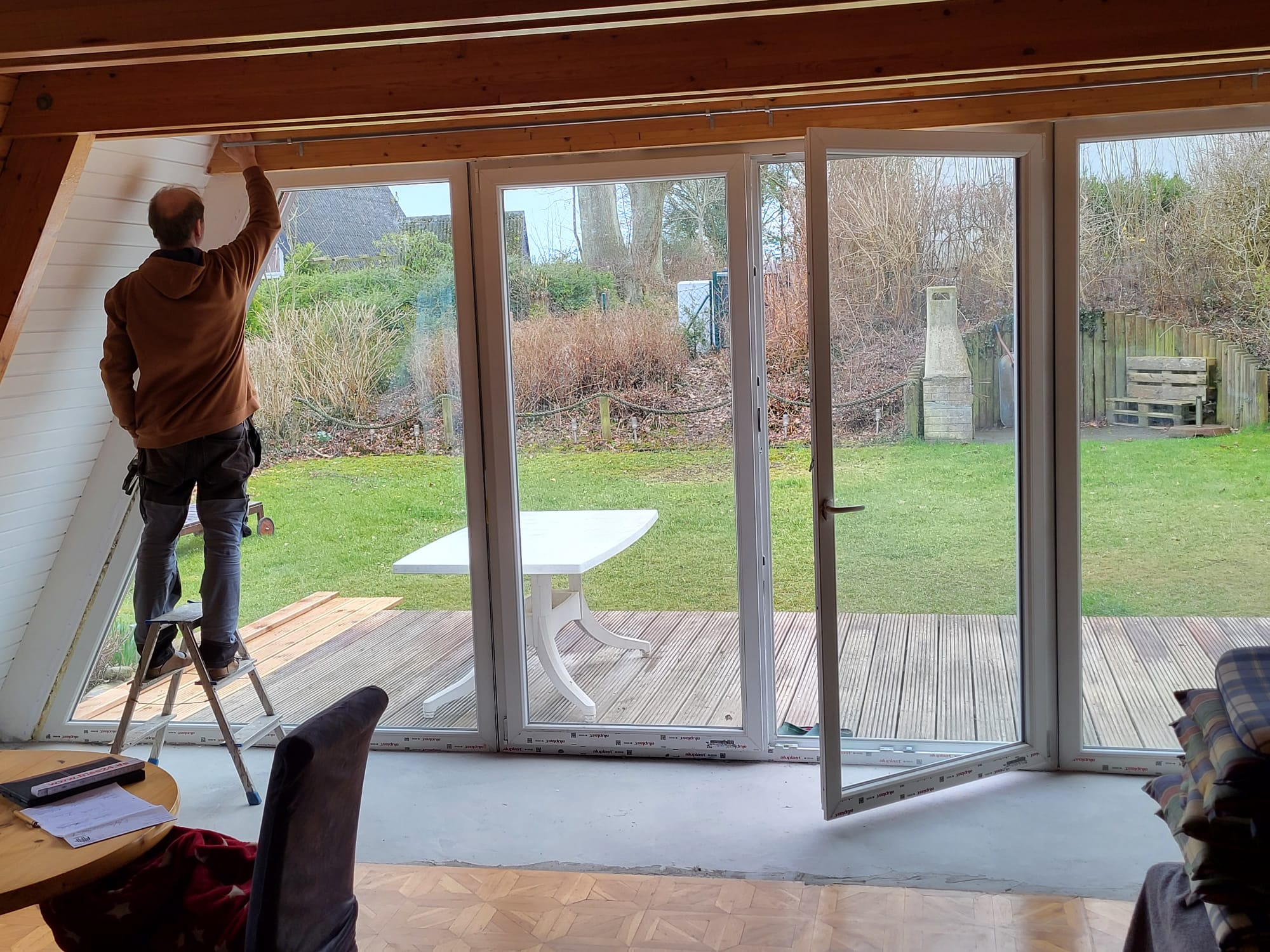 Person auf Leiter streicht Wand im Wohnzimmer, große Fenster zeigen Garten mit Terrasse.