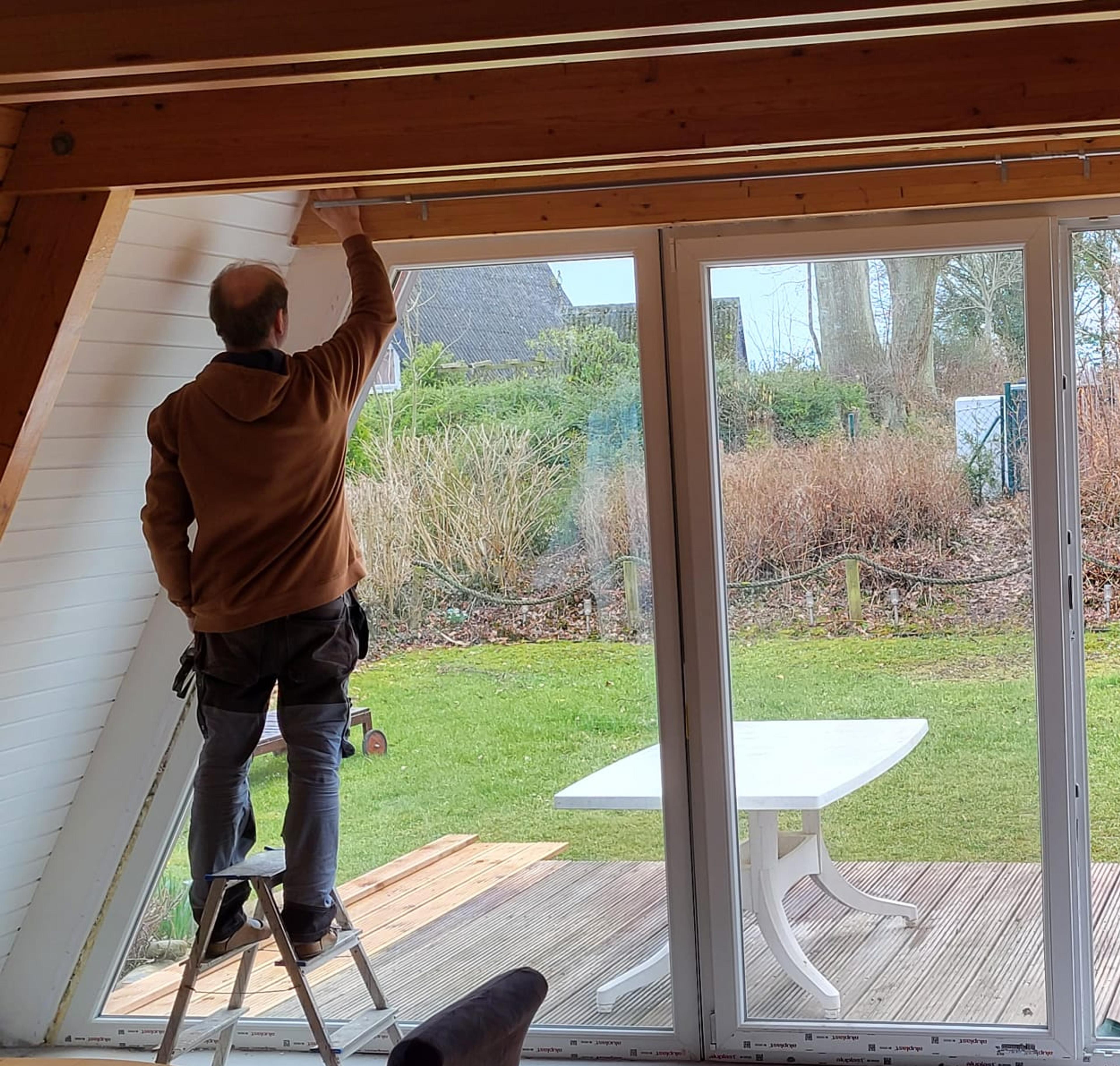 Person auf Leiter streicht Wand im Wohnzimmer, große Fenster zeigen Garten mit Terrasse.