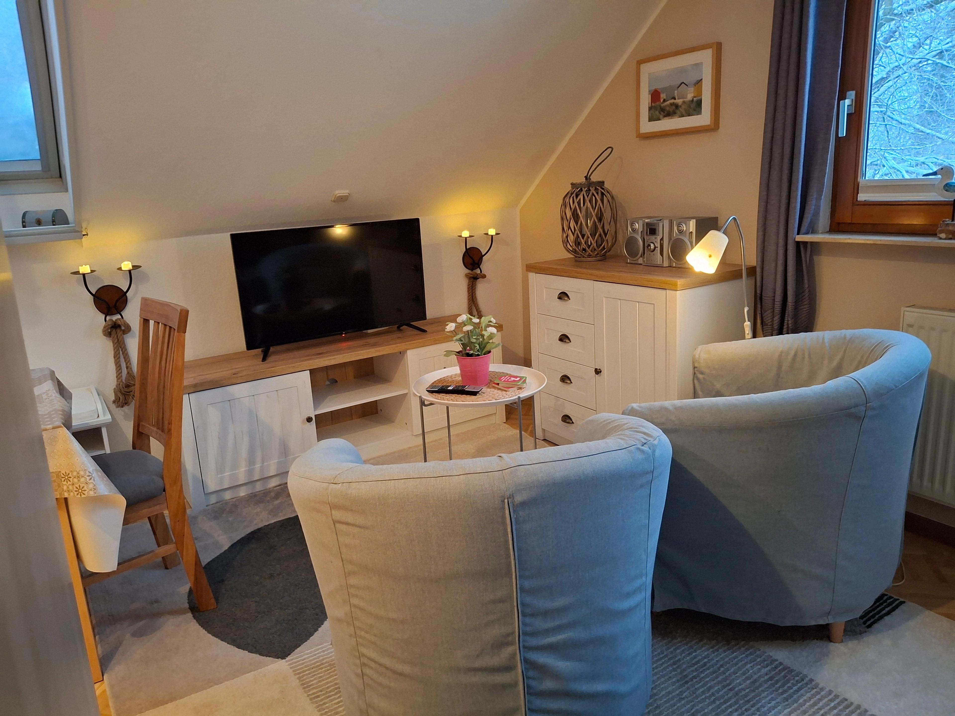 Wohnzimmer mit blauen Sesseln, Fernseher, Holzmöbeln und dezenter Beleuchtung.