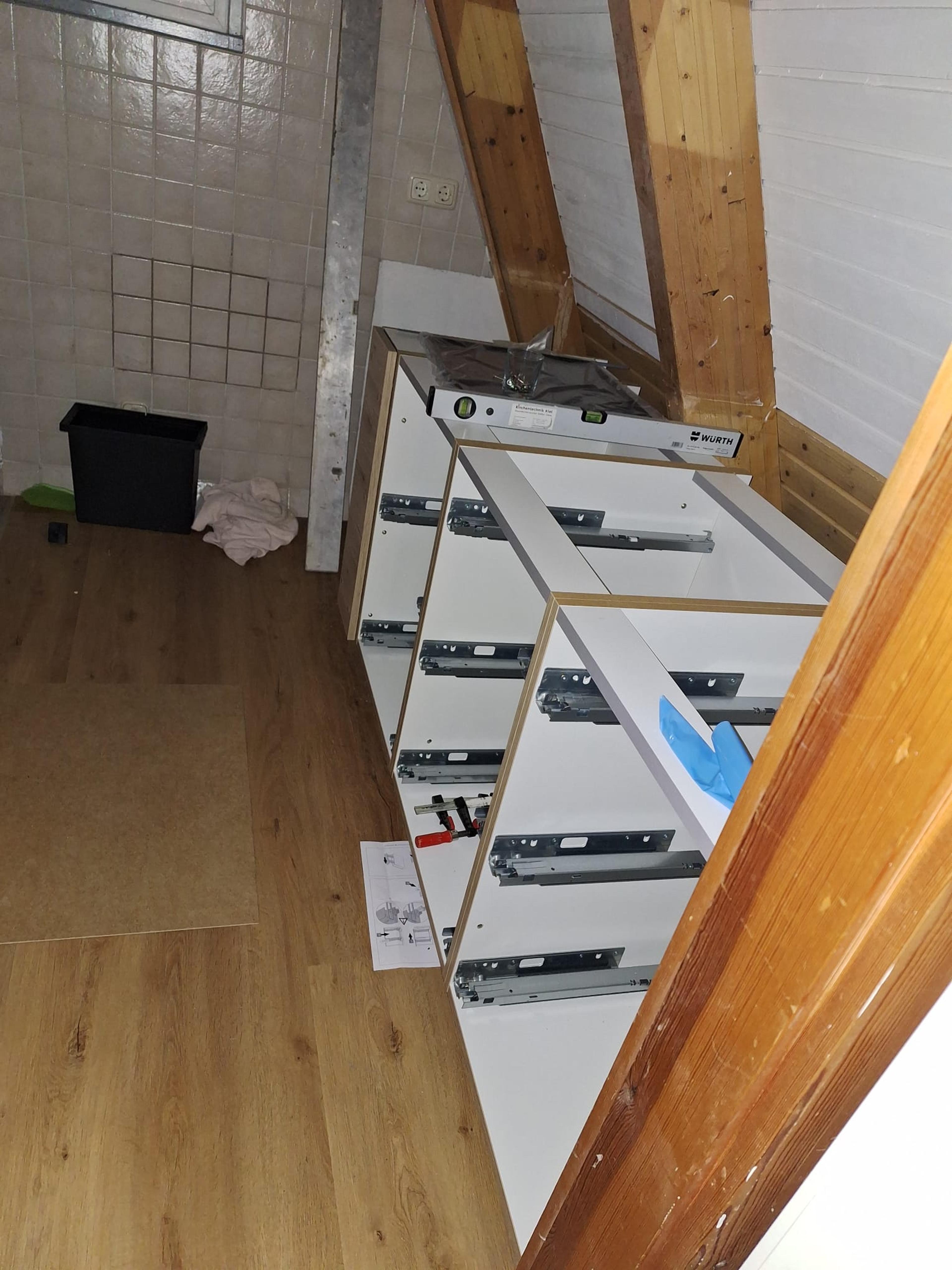 Küchenschränke im Aufbau in einem Dachgeschoss mit Holzverkleidung, Werkzeugen und Laminatboden.