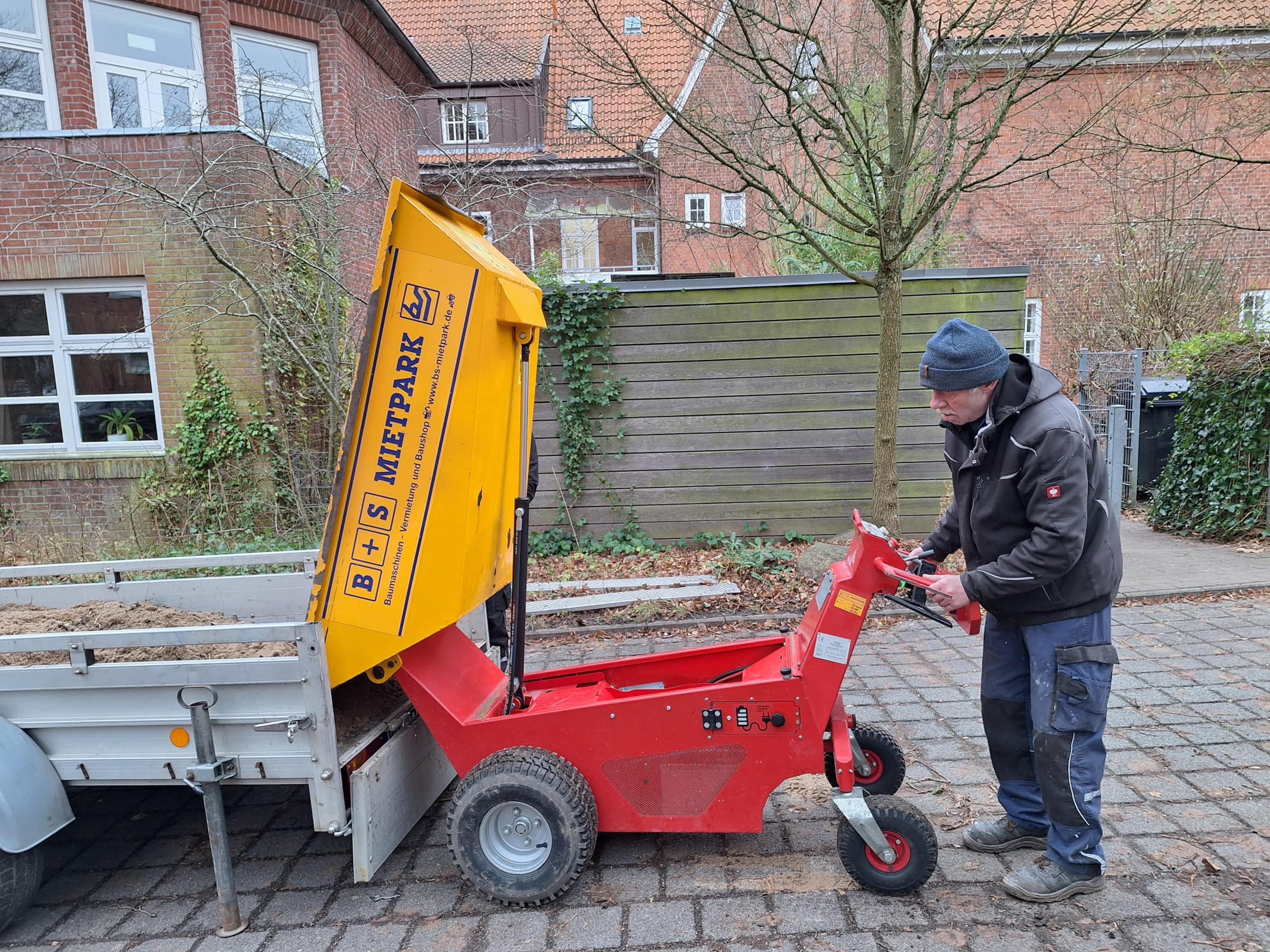 Arbeiter mit blauer Mütze bedient einen roten Mini-Dumper mit gelbem Kipper vor einem Ziegelhaus.