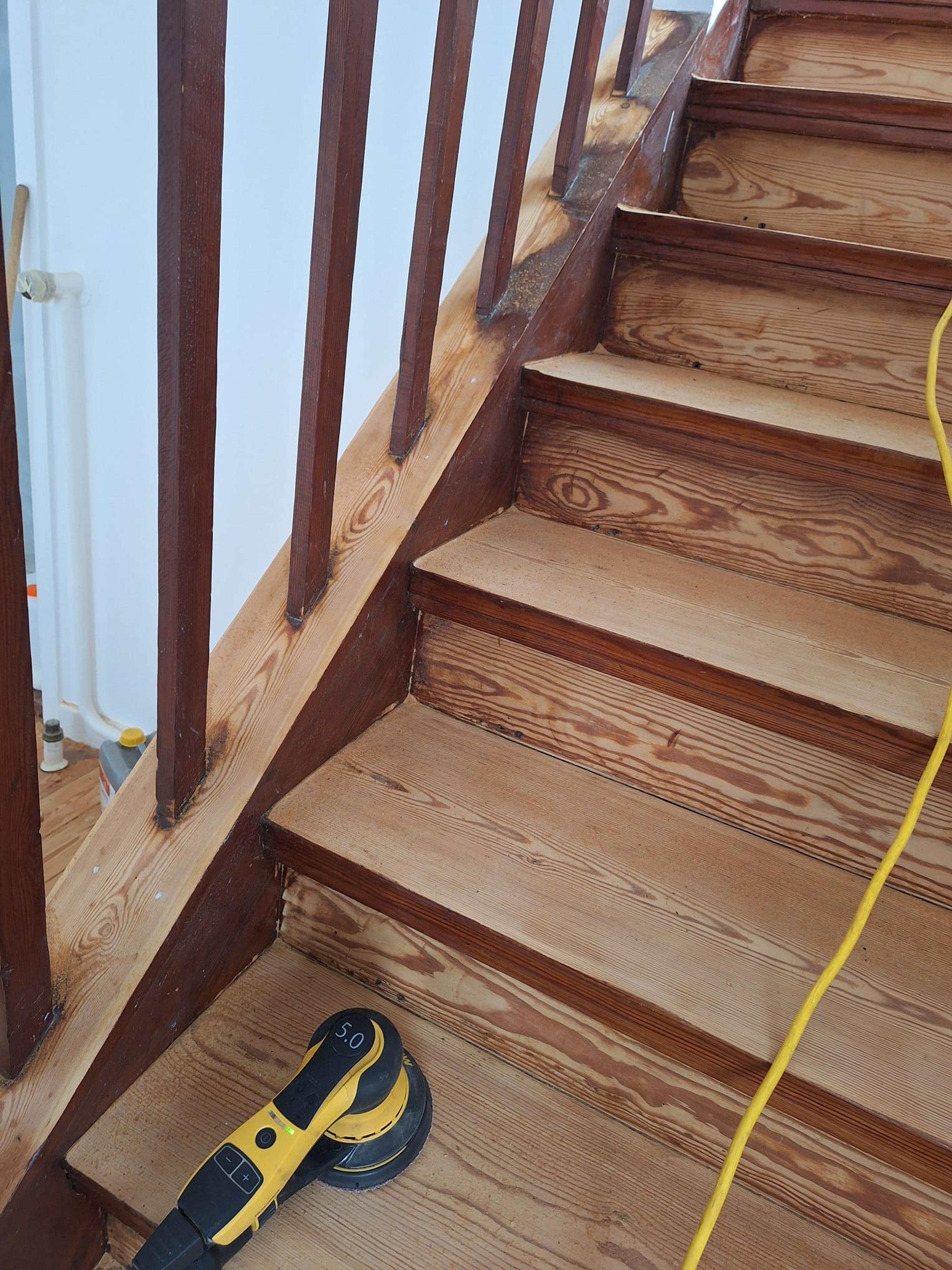 Holztreppe mit braunem Geländer, Schleifmaschine auf der Stufe, Holzmaserung sichtbar.