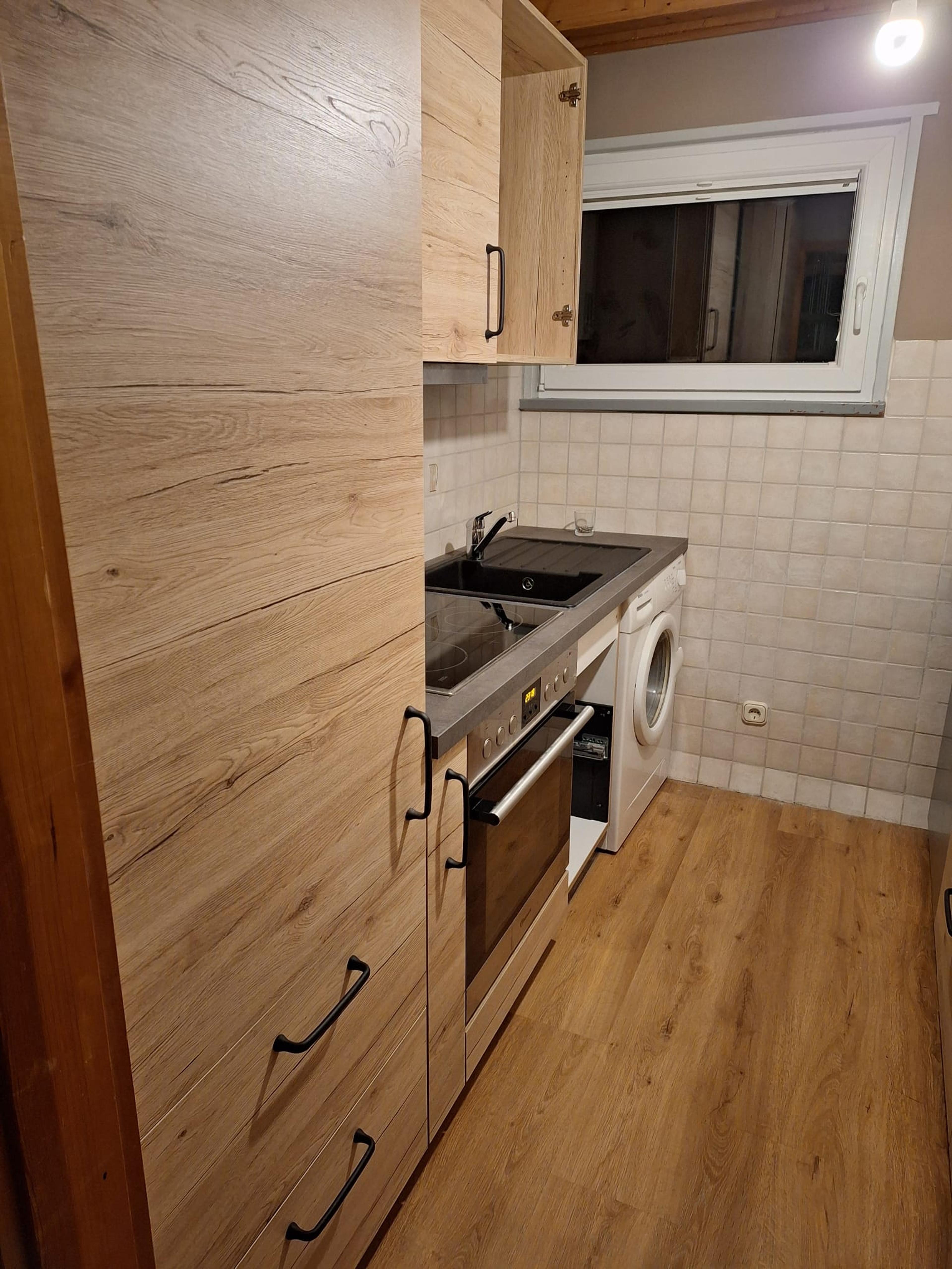 Kleine Küche mit Holzschränken, Einbauofen, und Waschmaschine in warmer Beleuchtung.