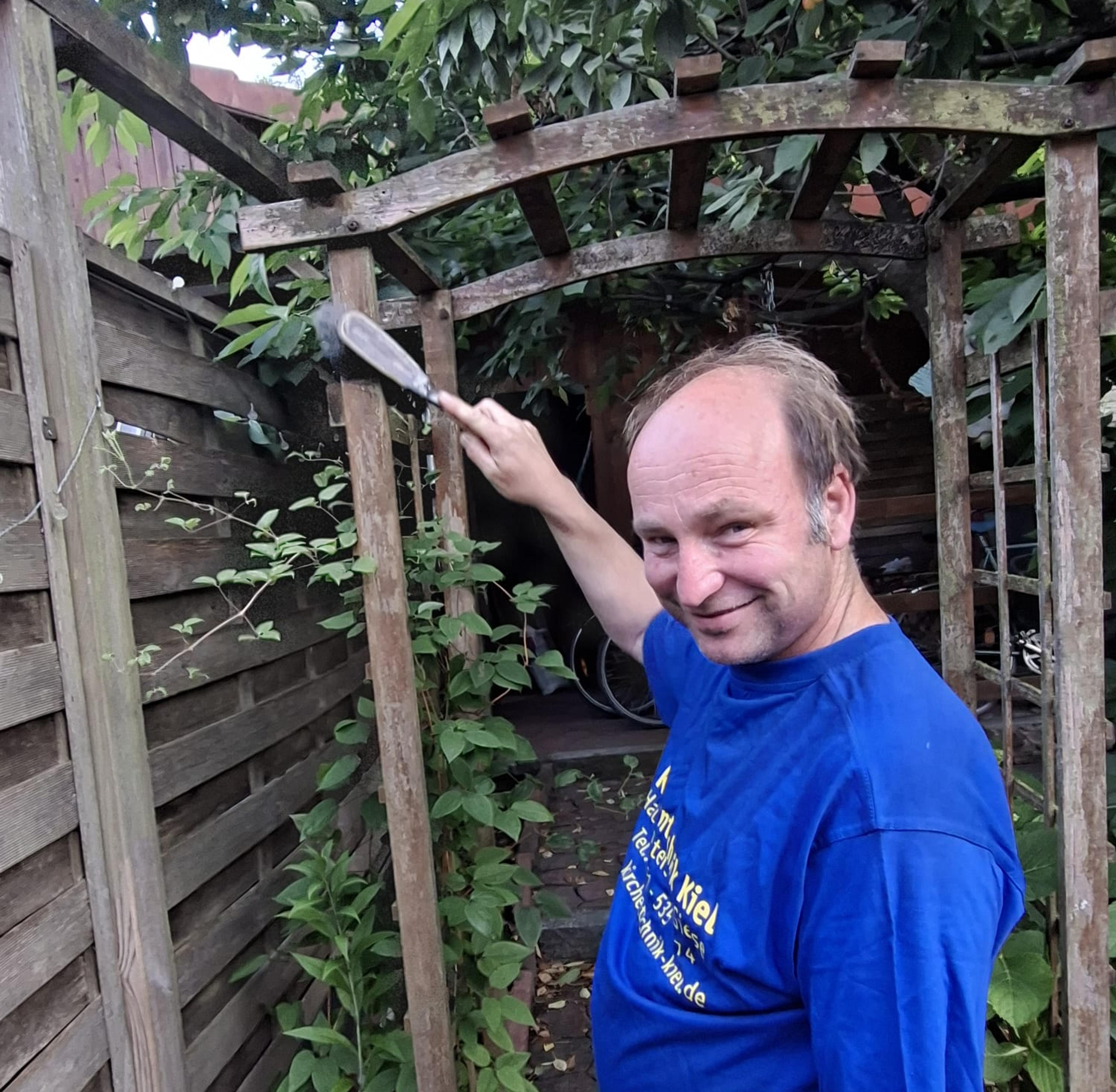Mann in blauem T-Shirt steht unter einem hölzernen Gartenbogen und pflegt Pflanzen.