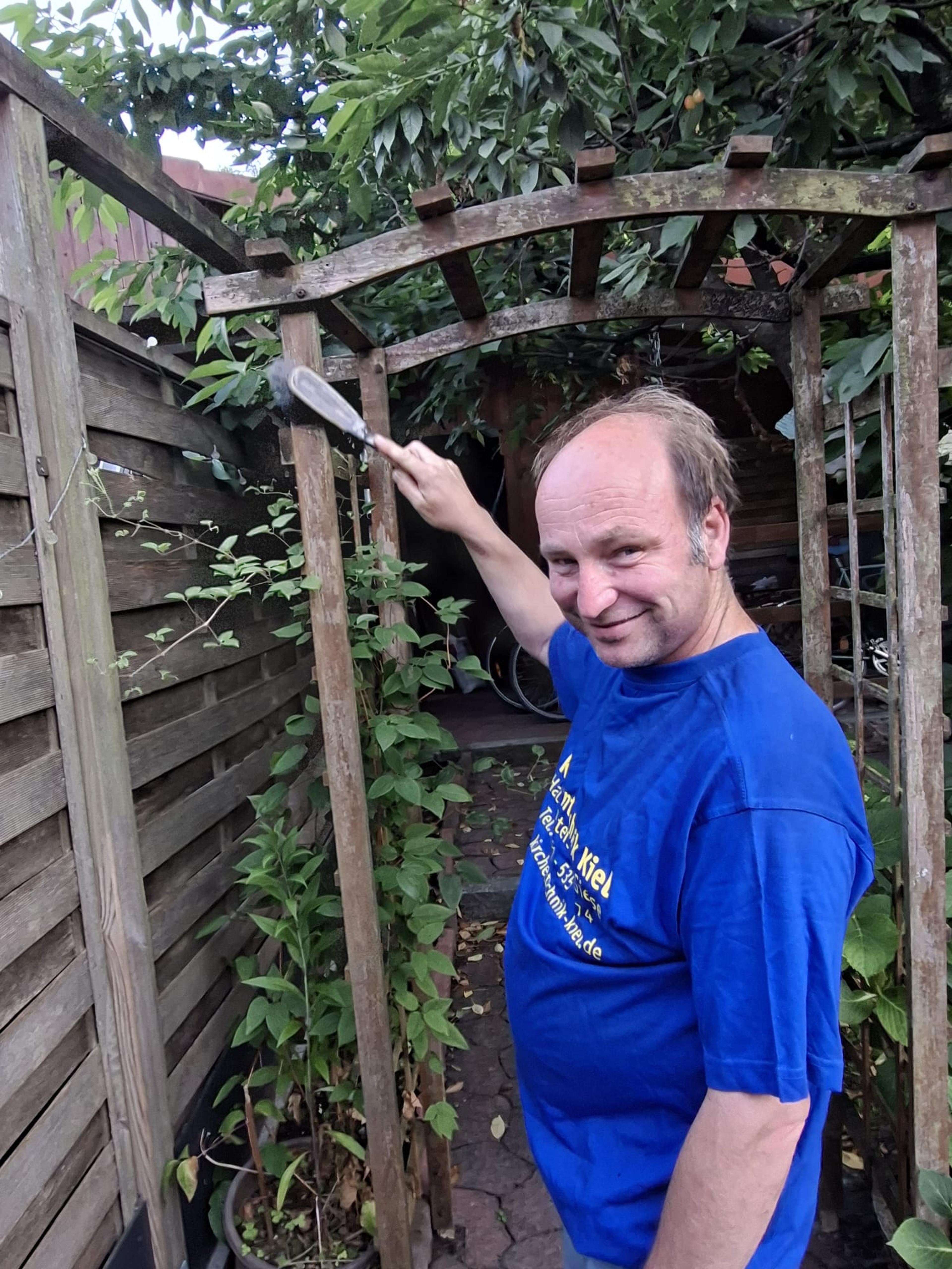 Mann in blauem T-Shirt pflegt Pflanzen unter einer hölzernen Pergola im Garten.