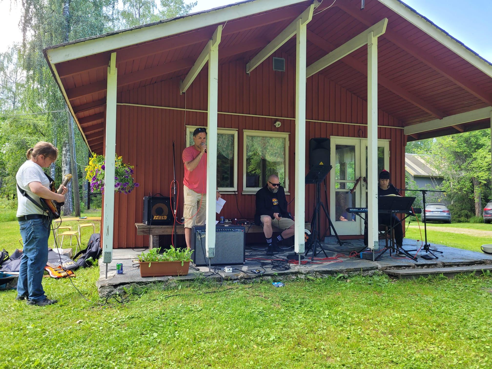 Bändi soittaa punaisen mökin katoksen alla kesäjuhlassa: basisti, laulaja, istuva laulaja ja kosketinsoittaja, 15.07.2023