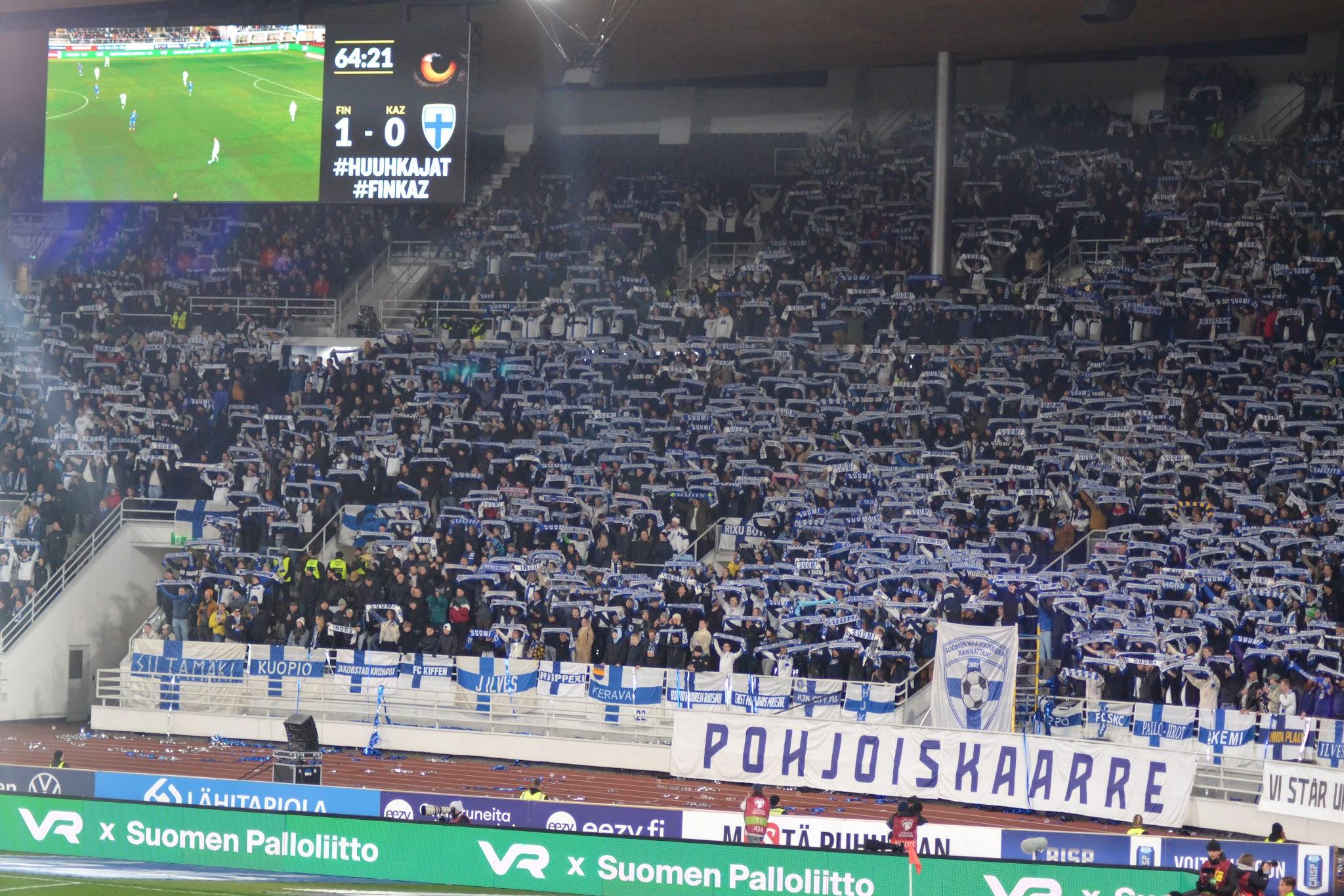 Pohjoiskaarren kannattajat kohottavat Suomi-kaulahuivejaan, tulostaululla FIN 1–0 KAZ Suomi–Kazakstan-ottelussa