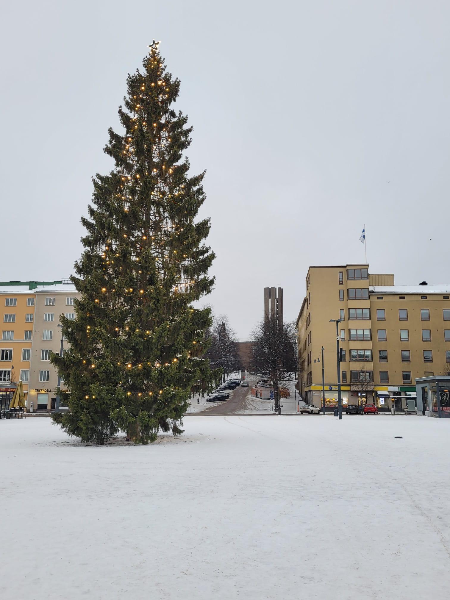 Jouluvaloin koristeltu kuusi lumisella Lahden torilla, taustalla kirkontorni ja katolla Suomen lippu, 06.12.2022