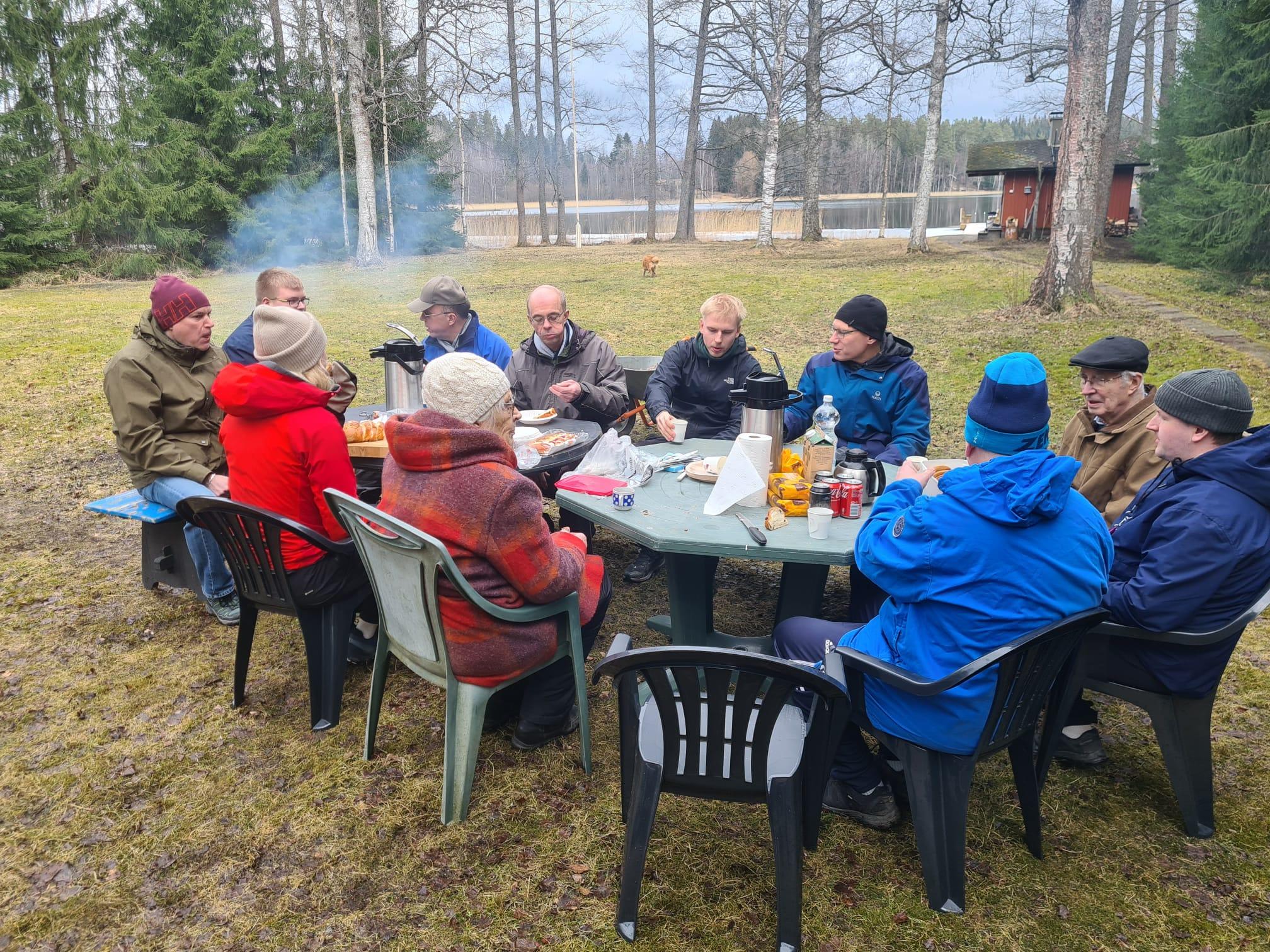 Pääsiäisen mölkyn kahvitauko: seurue istuu pöytien ääressä pihalla järven rannalla, 05.04.2026