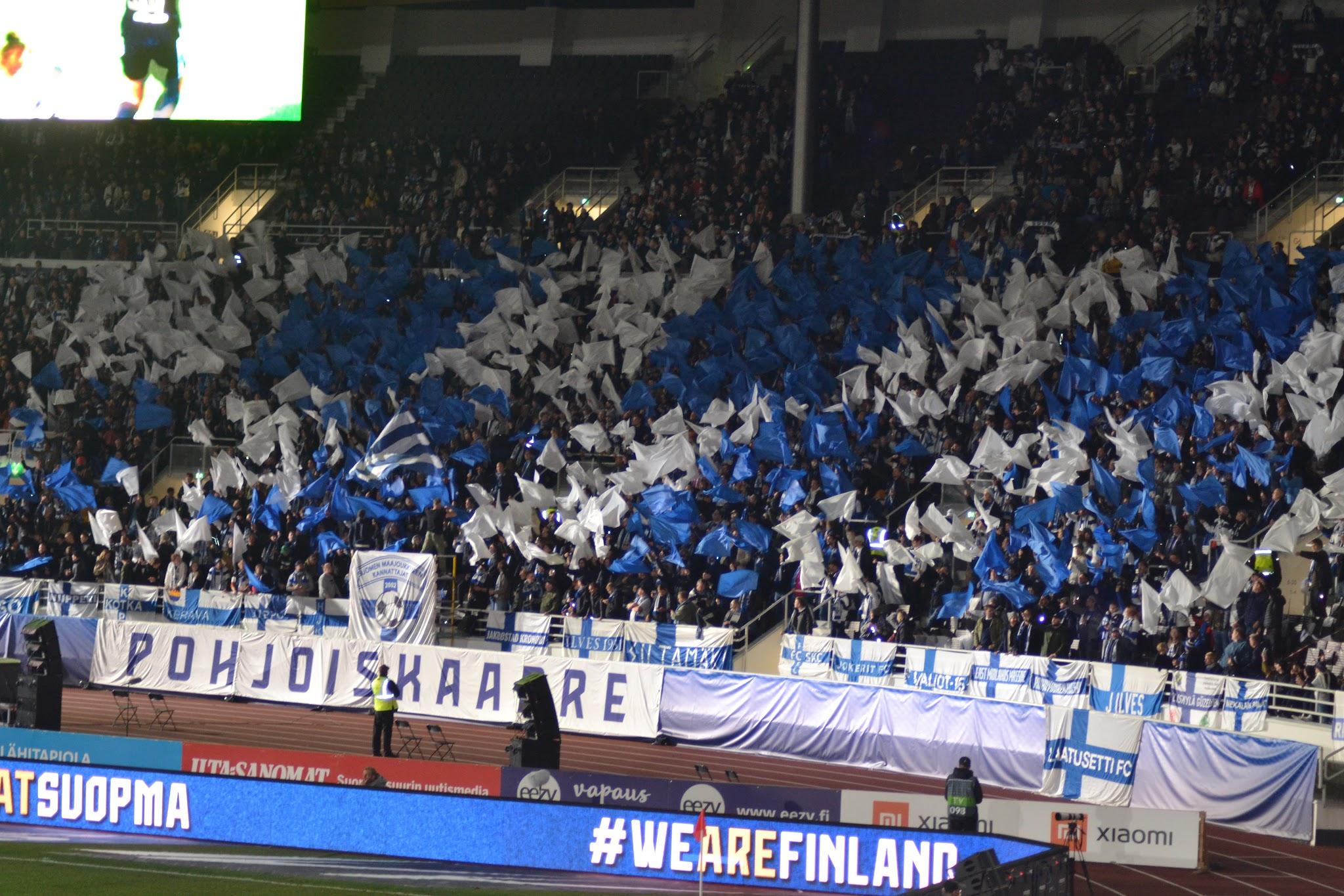 Suomen kannattajat heiluttavat sinisiä ja valkoisia lippuja Pohjoiskaarre-katsomossa, mainostaululla #WEAREFINLAND