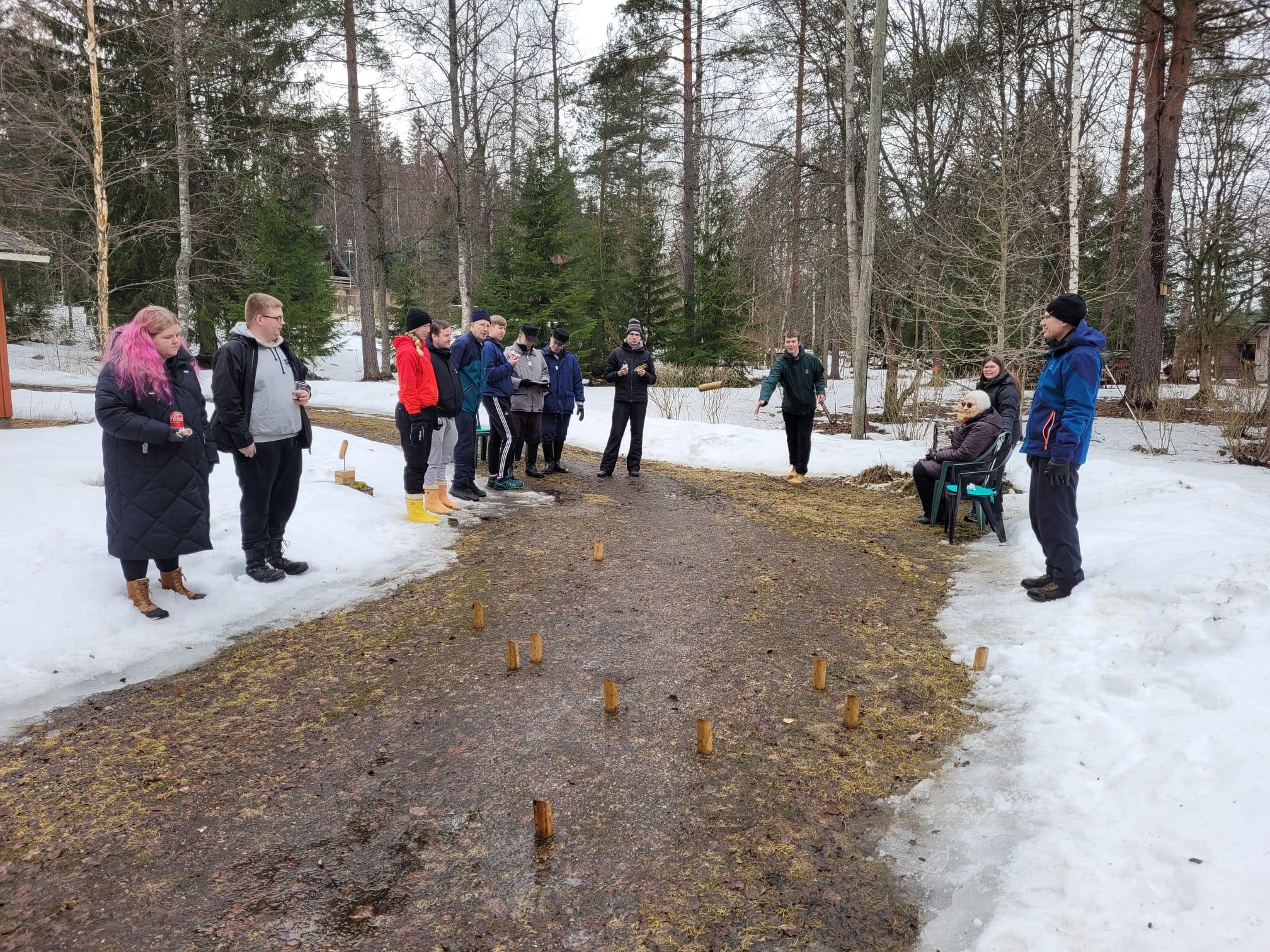 Pääsiäisen mölkkypeli käynnissä sulaneella hiekkatiellä lumen keskellä, pelaajat ja katsojat ympärillä, 31.03.2024