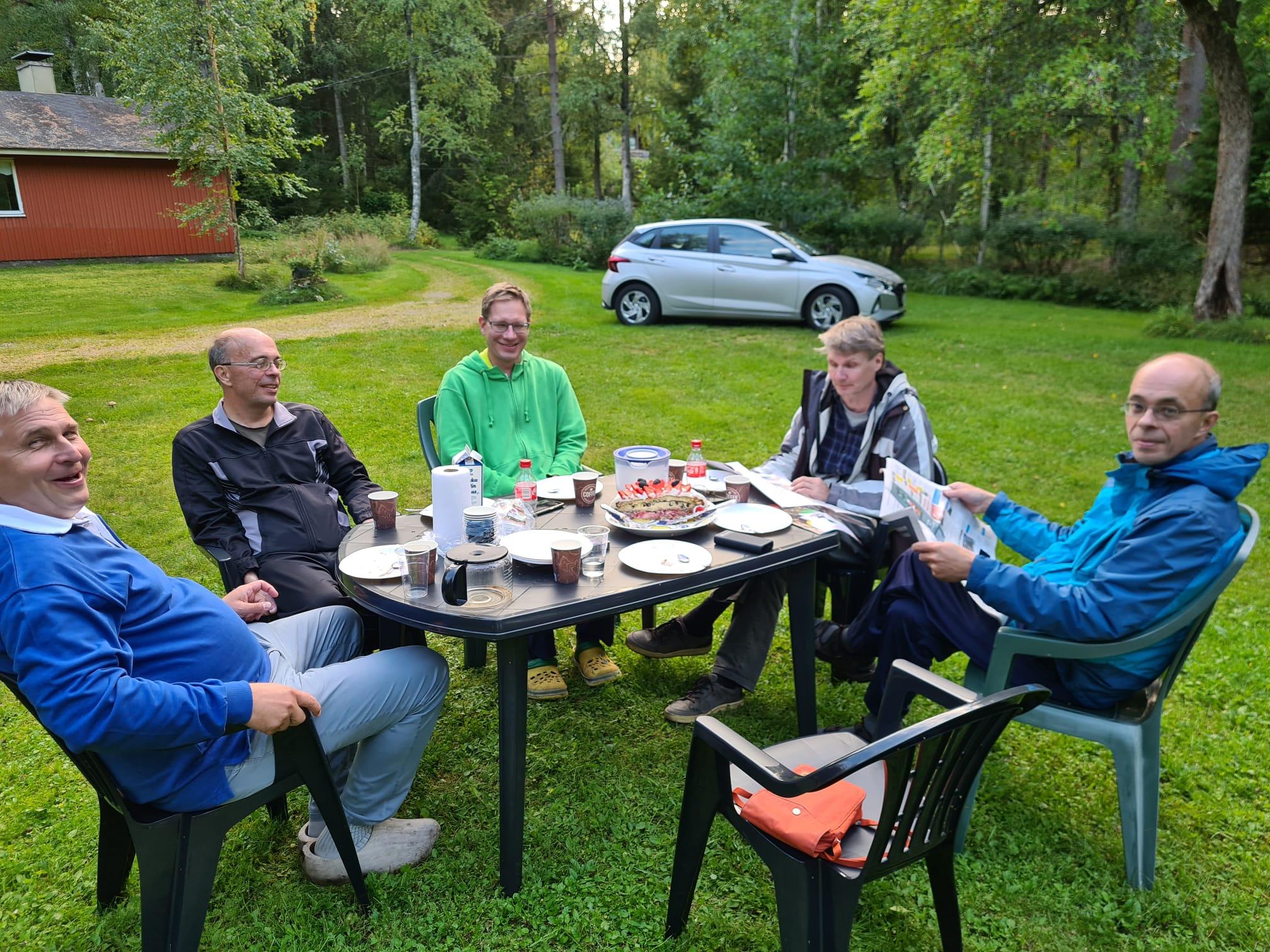 Viisi klubilaista kahvipöydän ääressä nurmikolla punaisen mökin pihalla, 26.08.2023