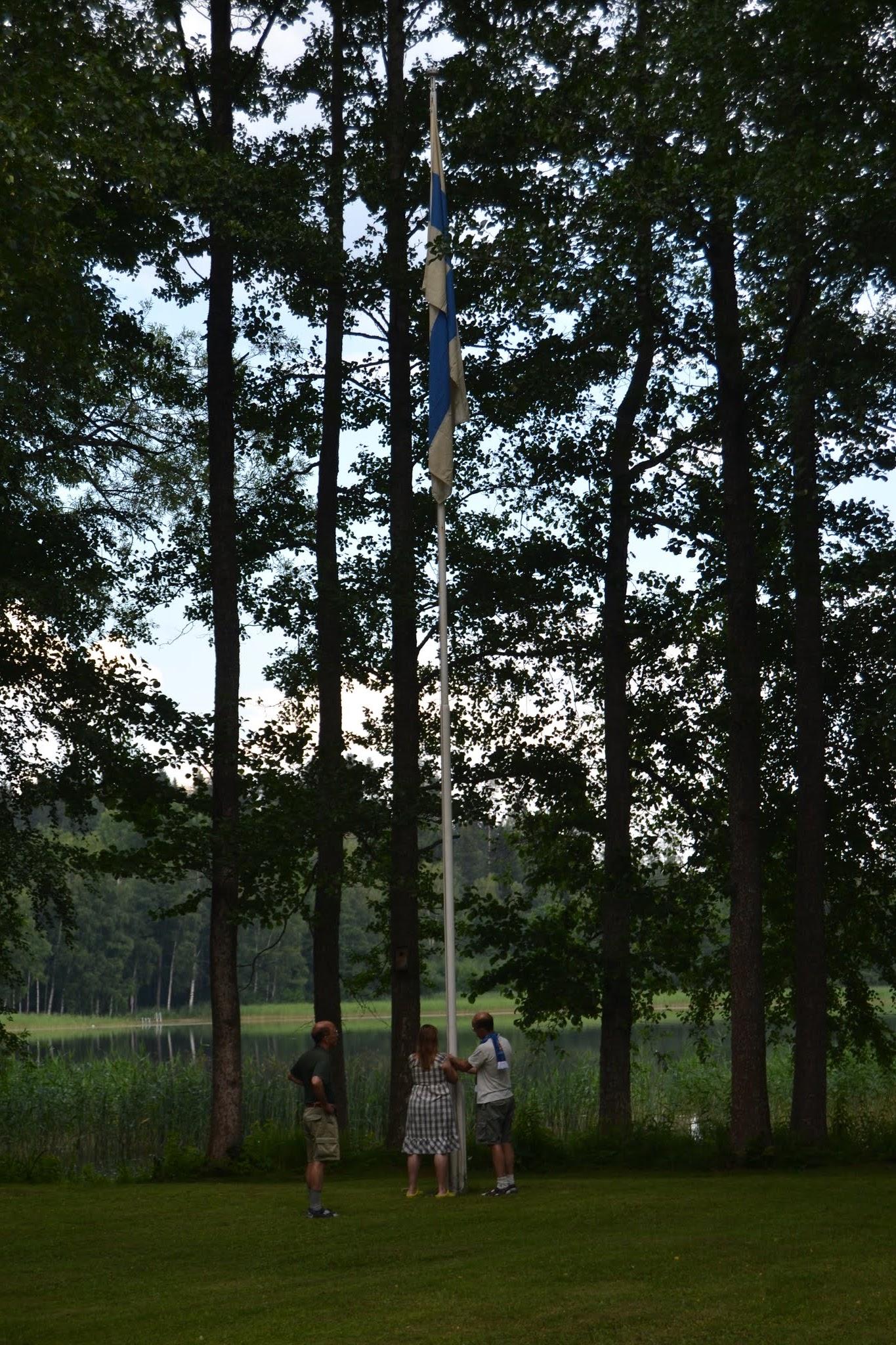 Kolme ihmistä nostaa Suomen lippua salkoon järvenrannan korkeiden puiden keskellä