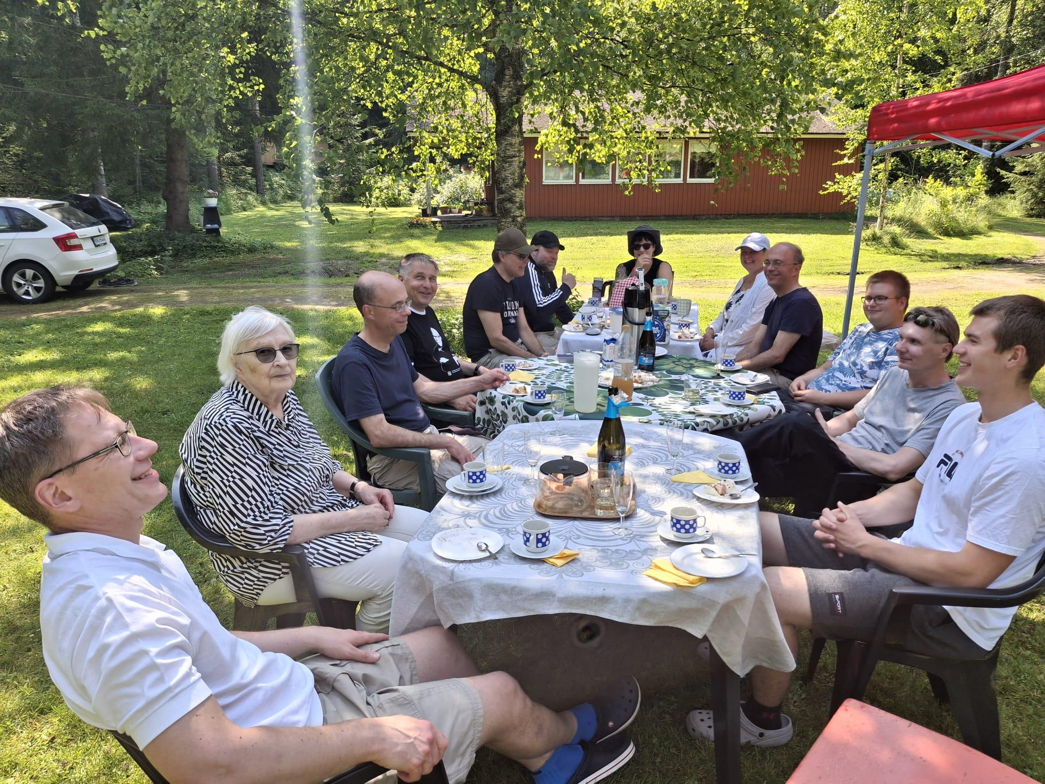 Klubilaisia kahvipöydän ääressä aurinkoisella pihalla punaisen talon edessä kesäjuhlassa, 12.07.2025