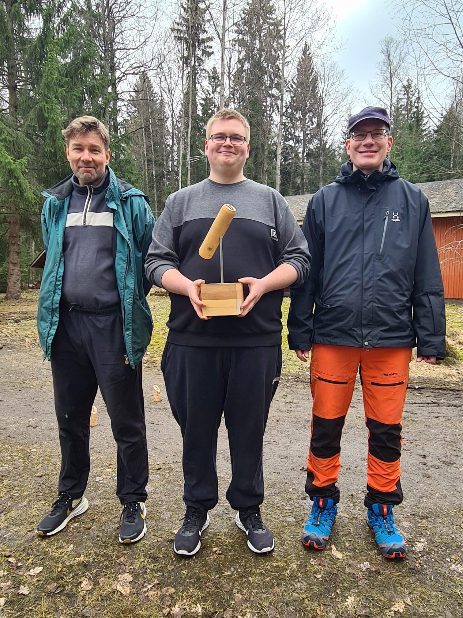 Mölkyn mitalistit: kolme miestä ulkona, keskimmäisellä puinen mölkkypalkinto käsissään, 18.04.2025