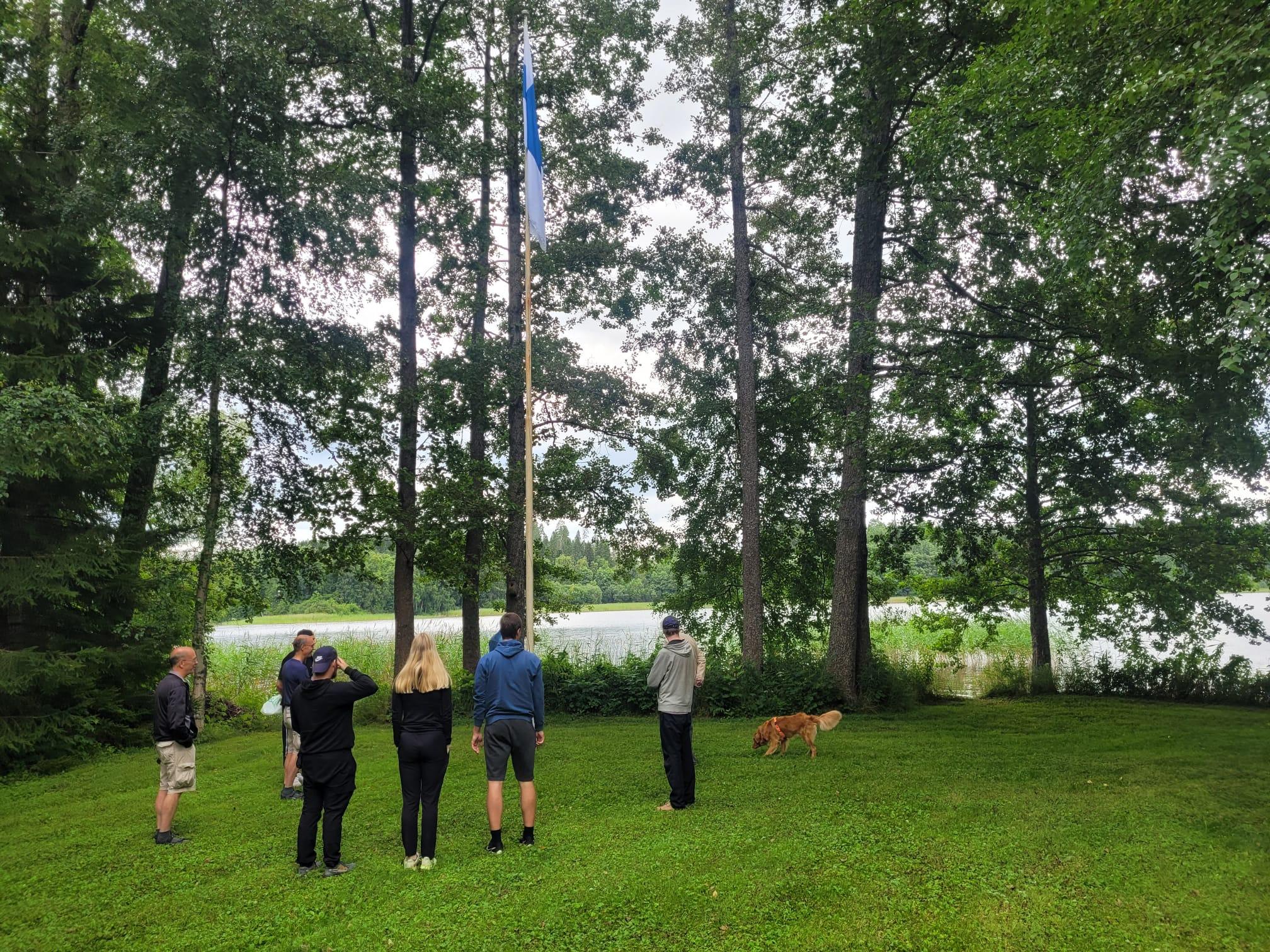 Seurue seuraa Suomen lipun nostoa salkoon järvenrantapihalla, vieressä koira, 25.07.2026
