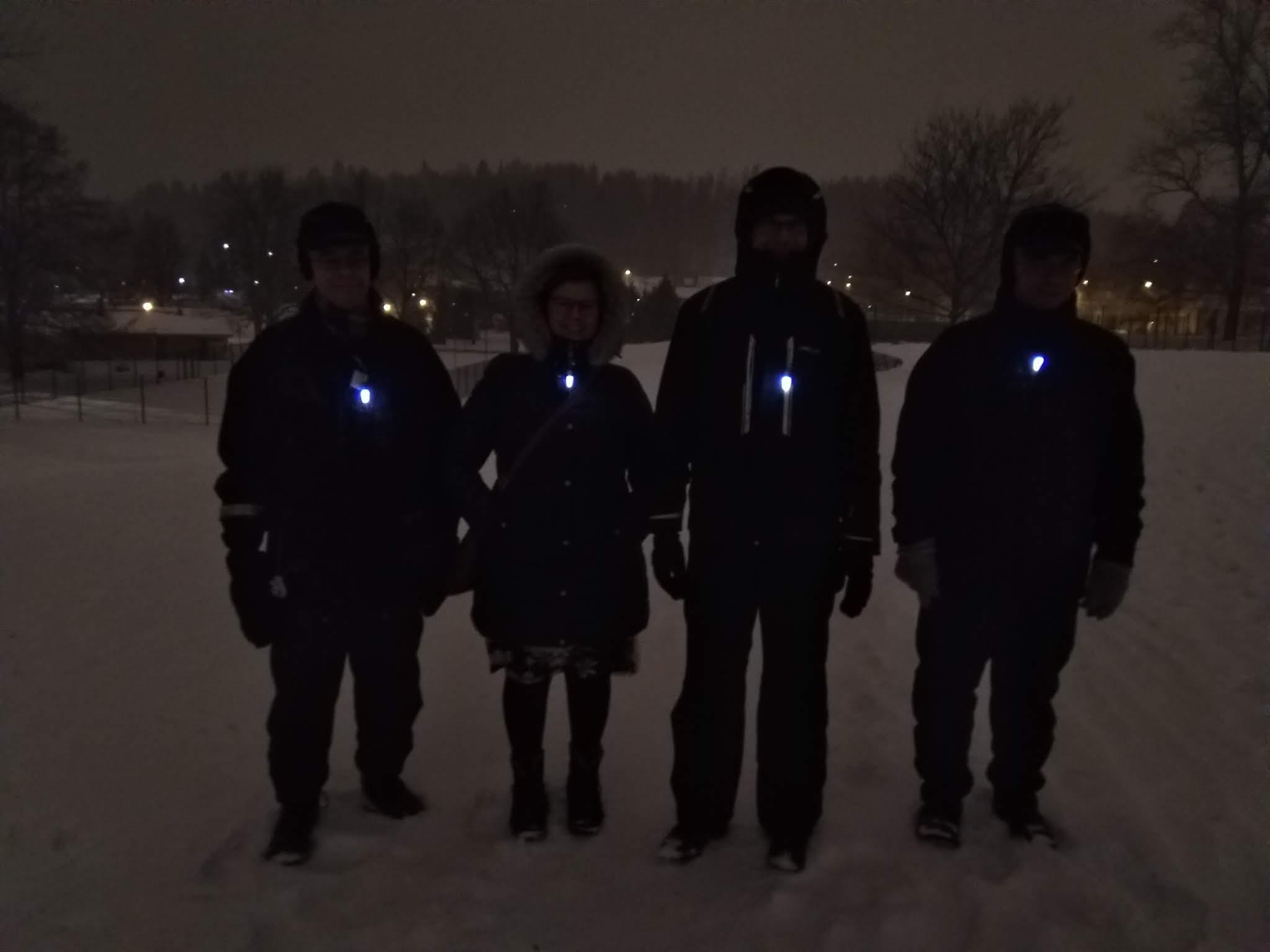 Neljä talvivaatteisiin pukeutunutta klubilaista lumisessa Kisapuistossa yöllä, rinnassa valoheijastimet, 31.12.2018