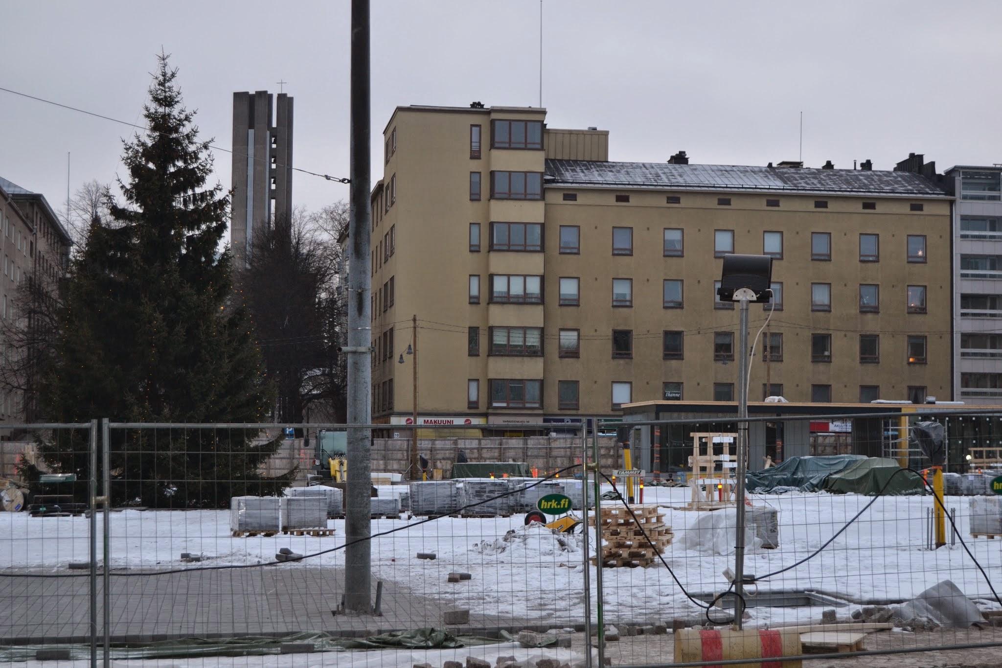 Joulukuusi työmaa-aitojen takana rakenteilla olevalla lumisella torilla Lahdessa, taustalla kirkontorni