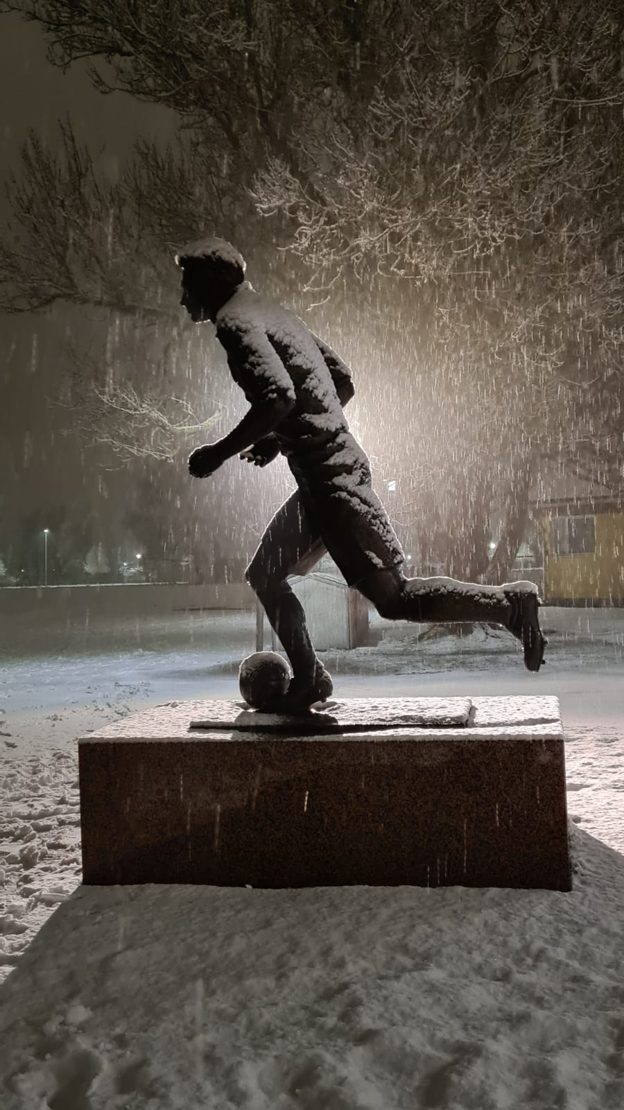 Lumen peittämä Litmasen patsas lumisateessa yöllä, valo loistaa patsaan takaa