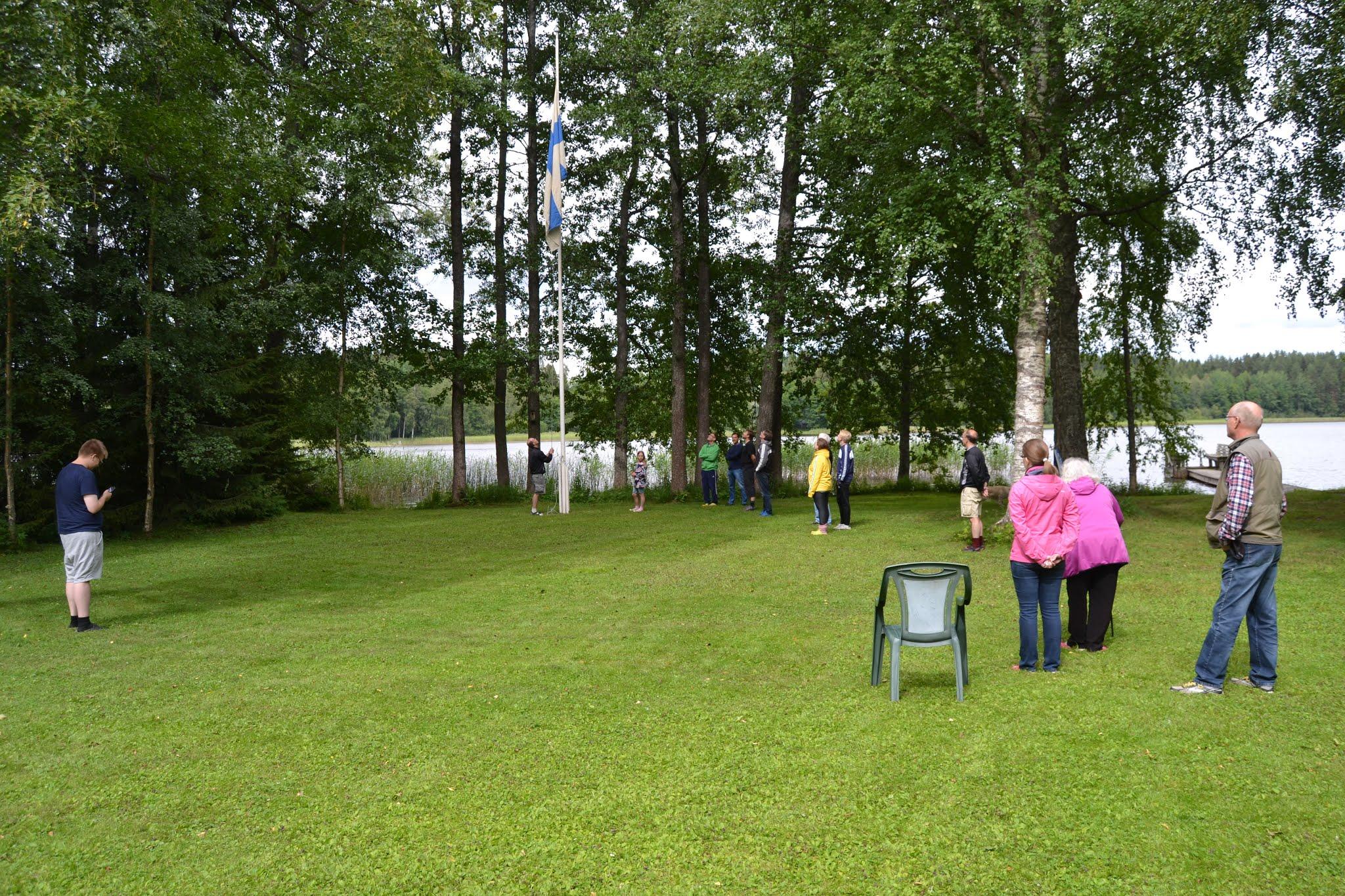Kesäjuhlan väki seuraa Suomen lipun nostoa järvenrantaisella nurmikolla koivujen keskellä