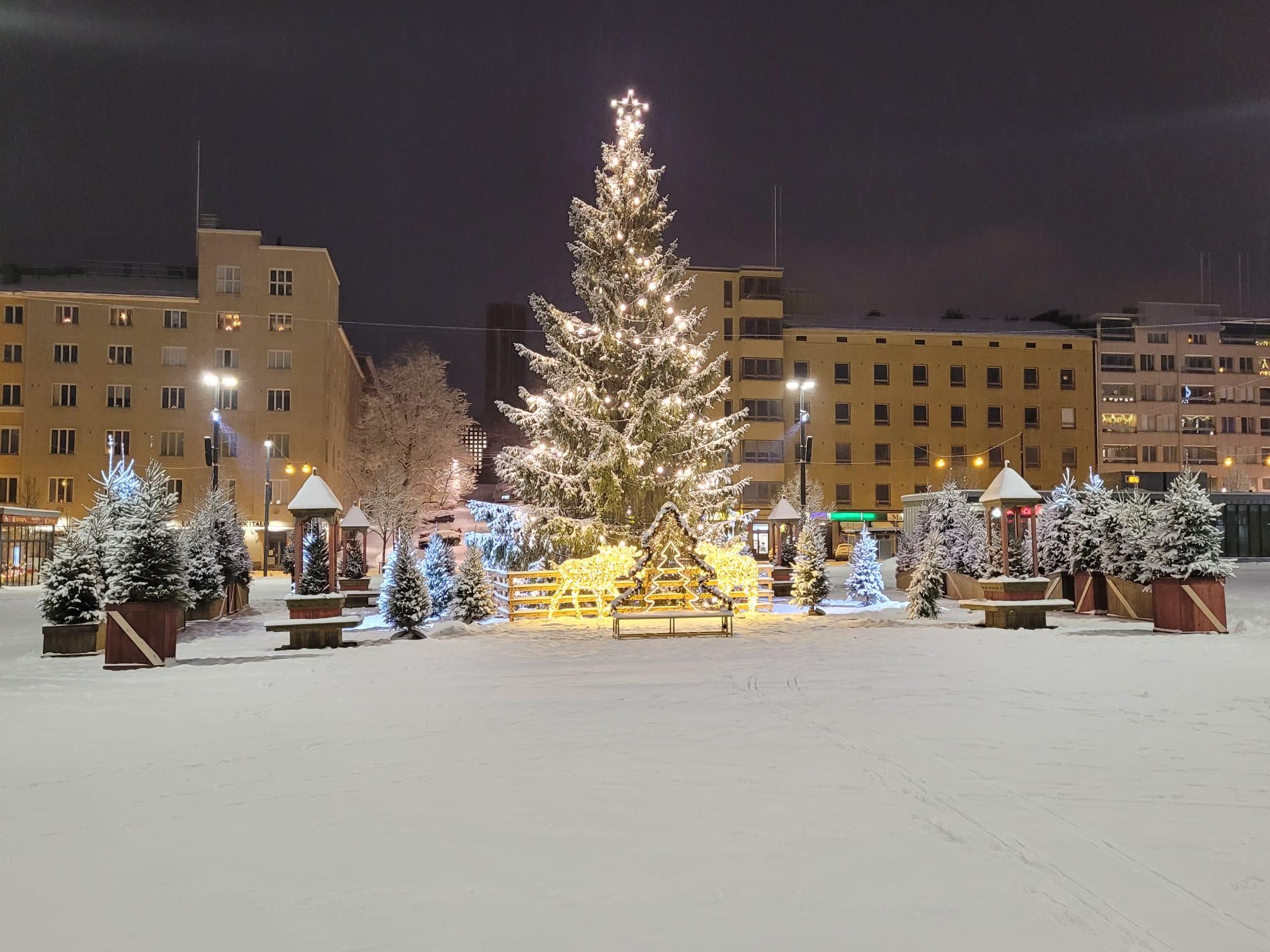 Lumen peittämä valaistu joulukuusi, valoporot ja pienet kuuset lumisella Lahden torilla pimeällä, 24.12.2023