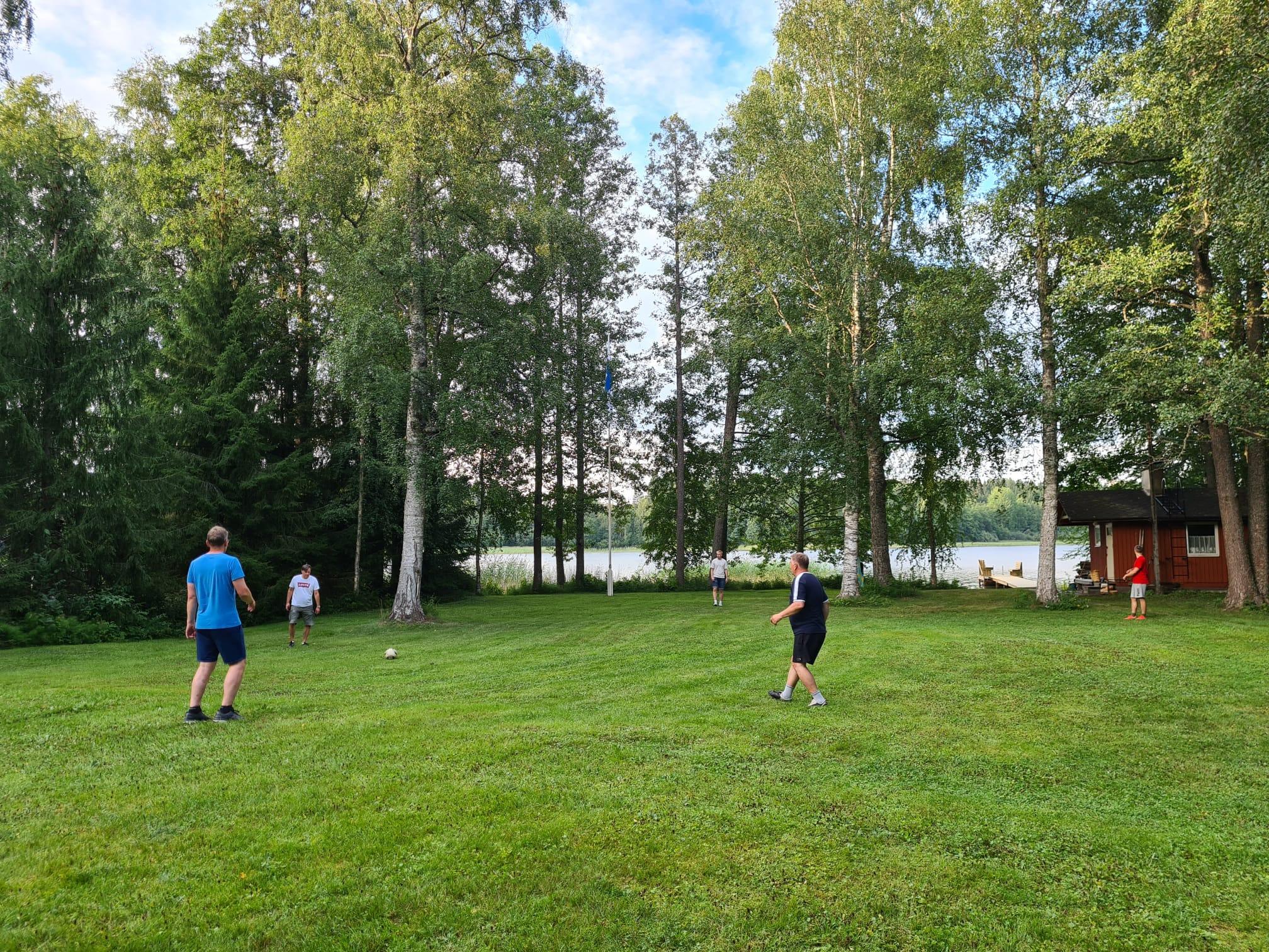 Klubilaiset pelaavat jalkapalloa nurmikentällä järvenrannassa punaisen saunan vieressä, 27.07.2024