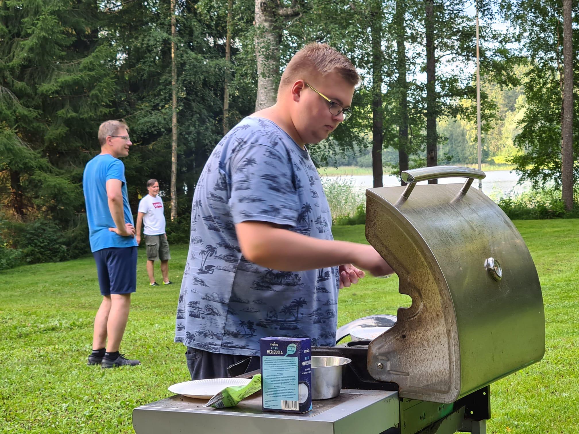 Nuori mies grillaa kaasugrillillä kesäjuhlassa, taustalla kaksi miestä nurmella ja järvi, 27.07.2024