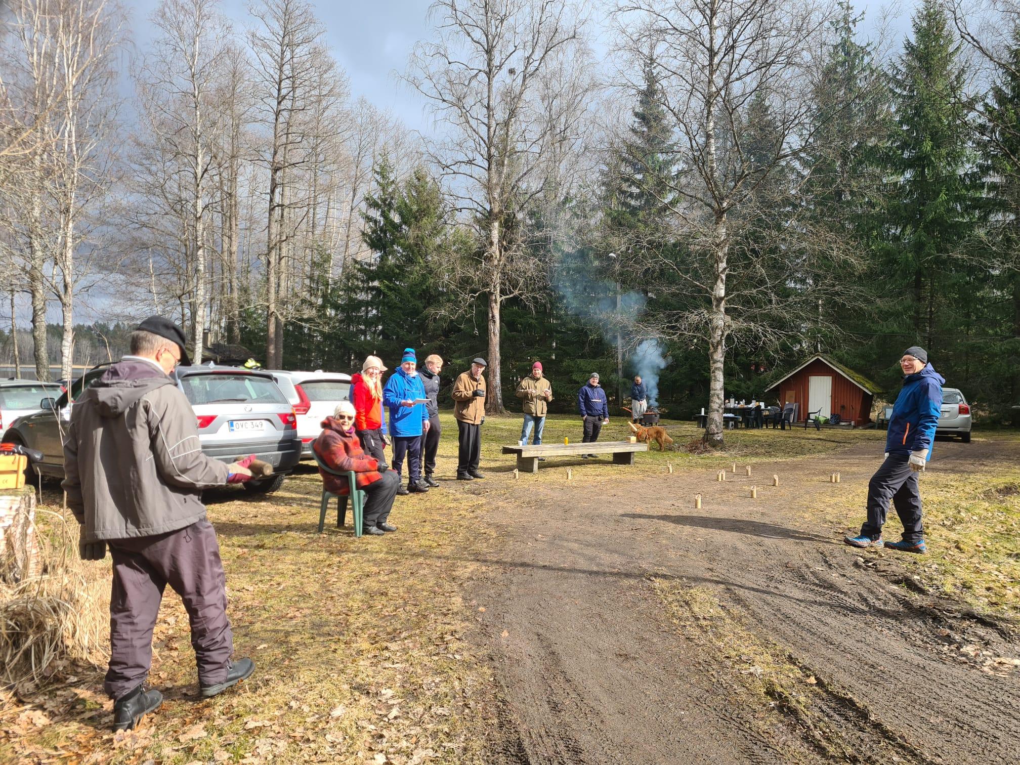 Mölkyn heittäjä valmistautuu heittoon, katsojat ja keilat järvenrantapihalla pääsiäisenä, 05.04.2026