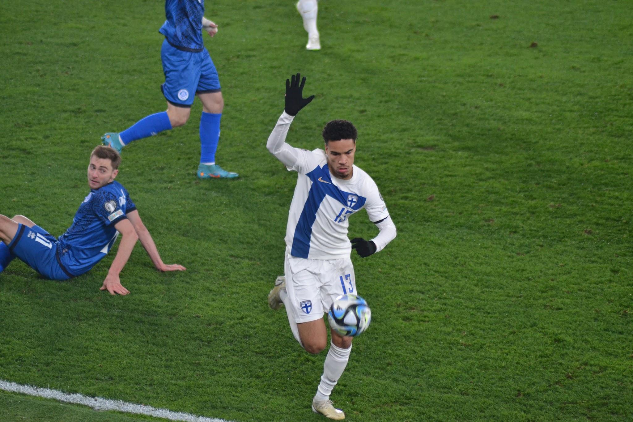 Suomen pelaaja numero 13 kuljettaa palloa, Kazakstanin pelaaja istuu nurmella