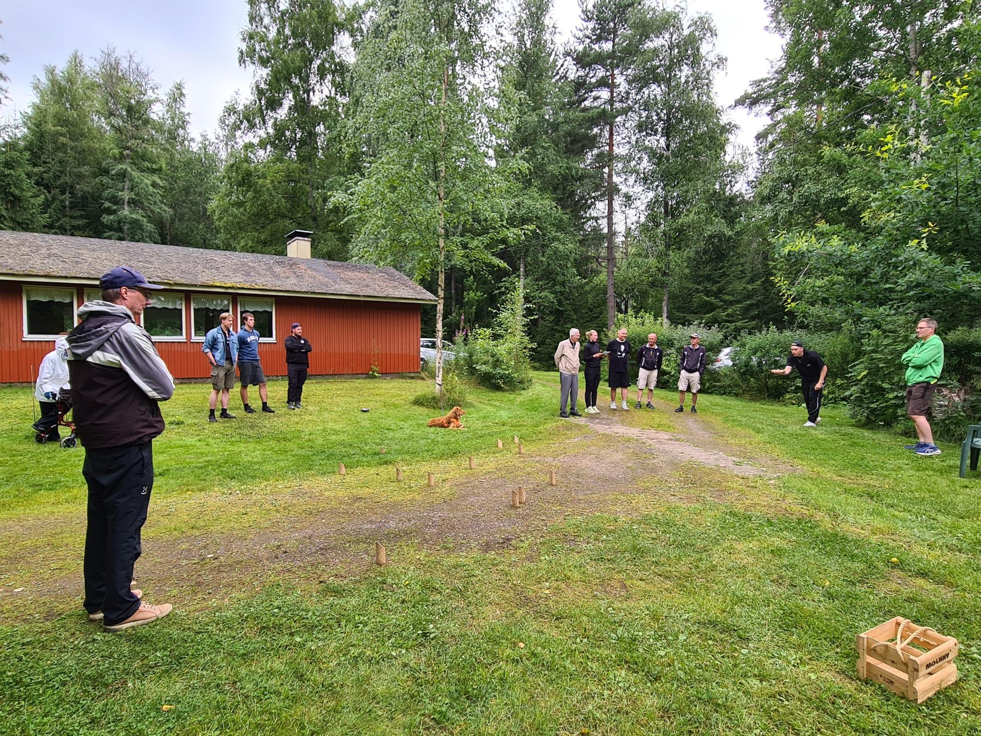 Mölkkypeli käynnissä nurmikolla punaisen mökin pihassa, pelaajat seuraavat heittoa, 25.07.2026