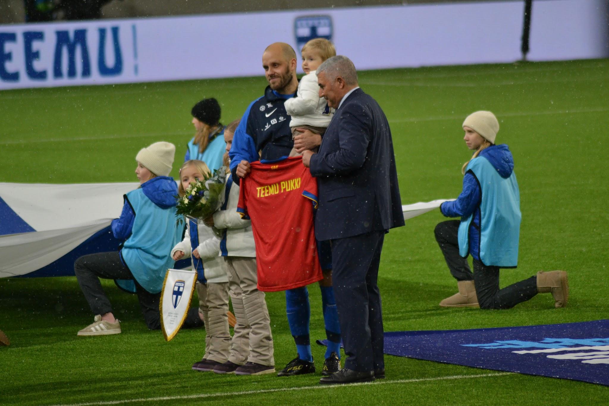 Kentällä esitellään punainen TEEMU PUKKI -nimikoitu pelipaita, taustalla TEEMU!-banderolli ja Suomen lippu