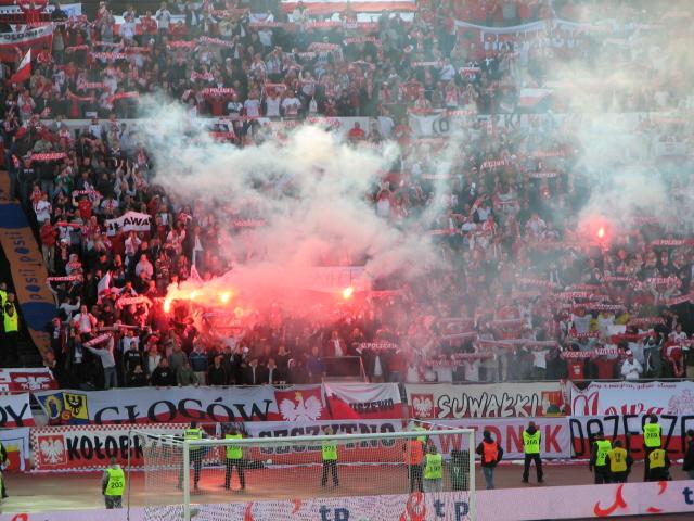 Punavalkoisiin pukeutuneet kannattajat polttavat soihtuja katsomossa, edessä Puolan kotkalla koristeltuja banderolleja, 12.09.2007