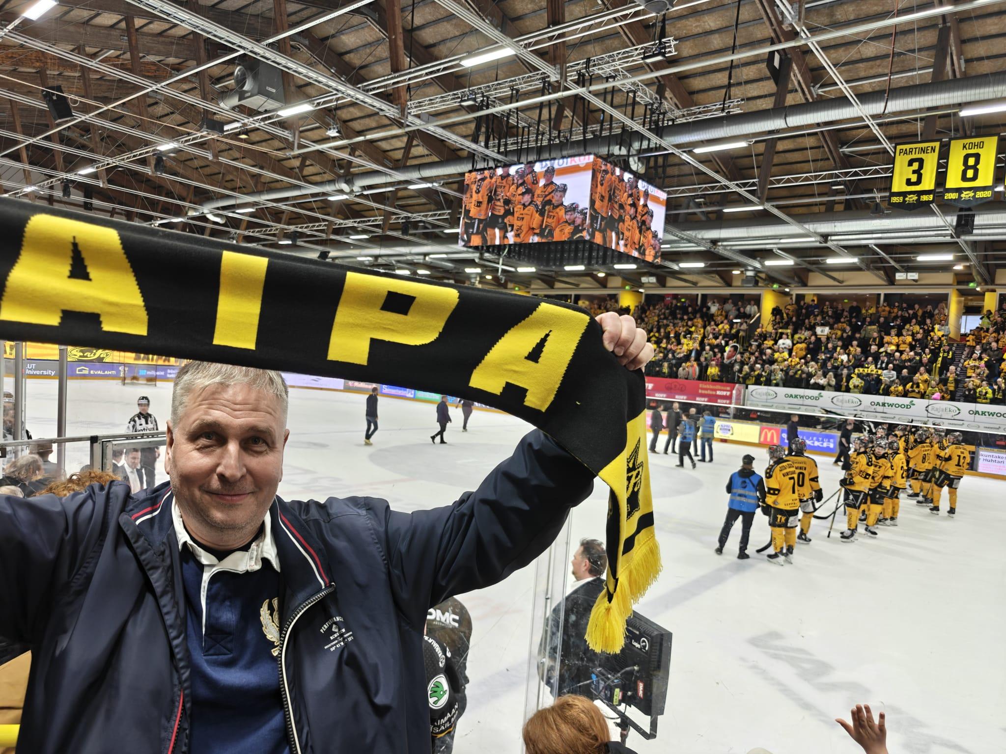 Simo kohottaa SaiPan kannattajahuivia jäähallissa, taustalla keltapaitaiset pelaajat jäällä, 30.04.2026