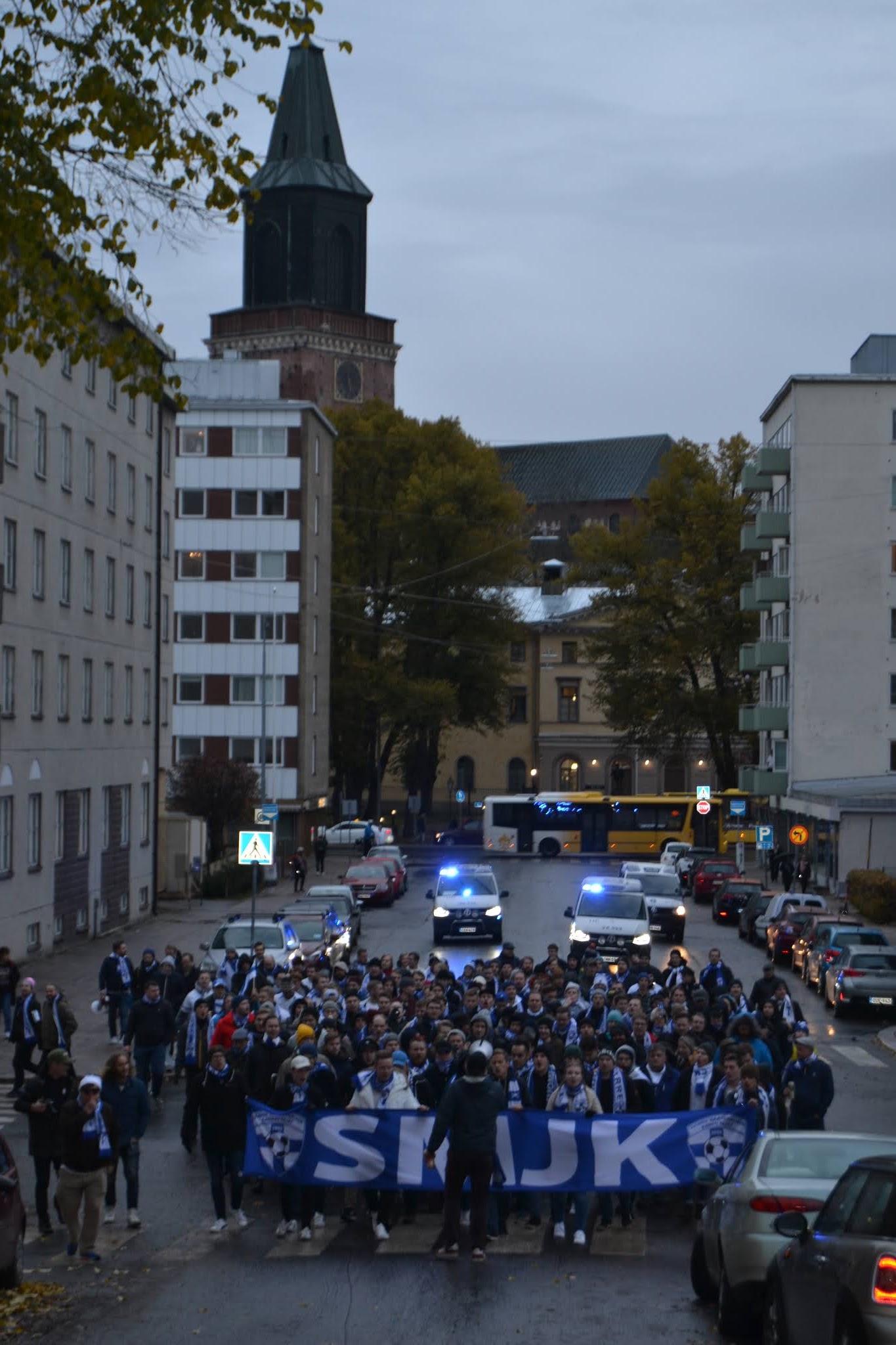 Suomen kannattajat marssivat sinivalkoisin kaulahuivein SMJK-banderollin takana Turun kadulla, taustalla kirkontorni ja poliisiautot