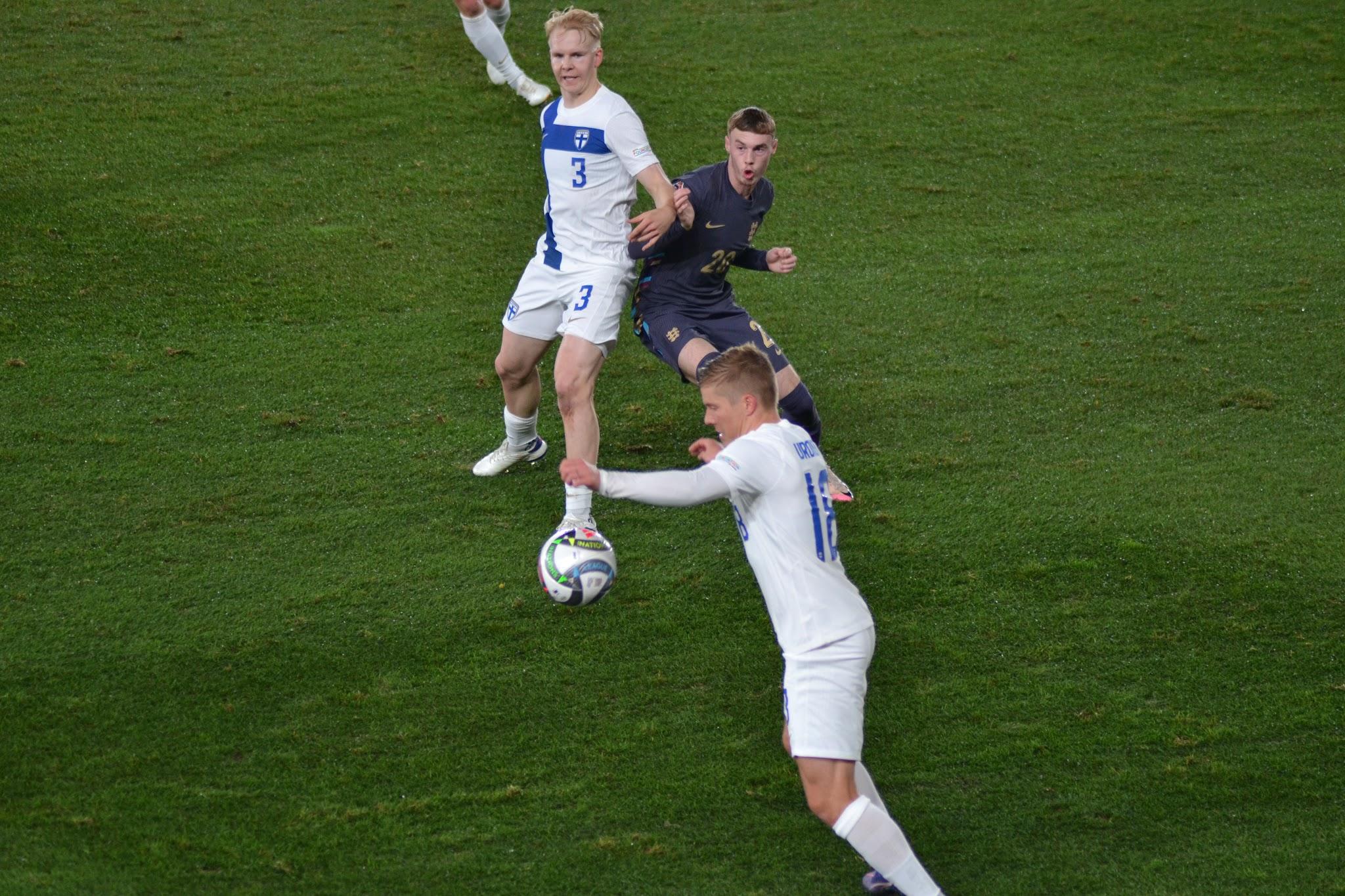 Suomi–Englanti-ottelussa Suomen valkopaitaiset numerot 3 ja 18 ja Englannin tummapaitainen pelaaja pallon luona