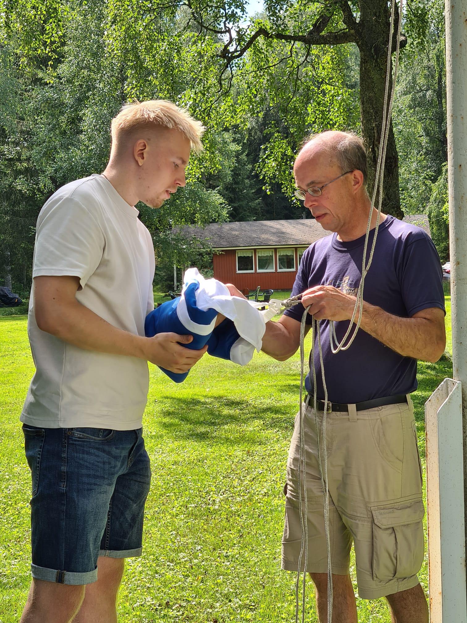 Kaksi miestä kiinnittää Suomen lippua salkoon kesäjuhlan lipunnostossa, 27.07.2024