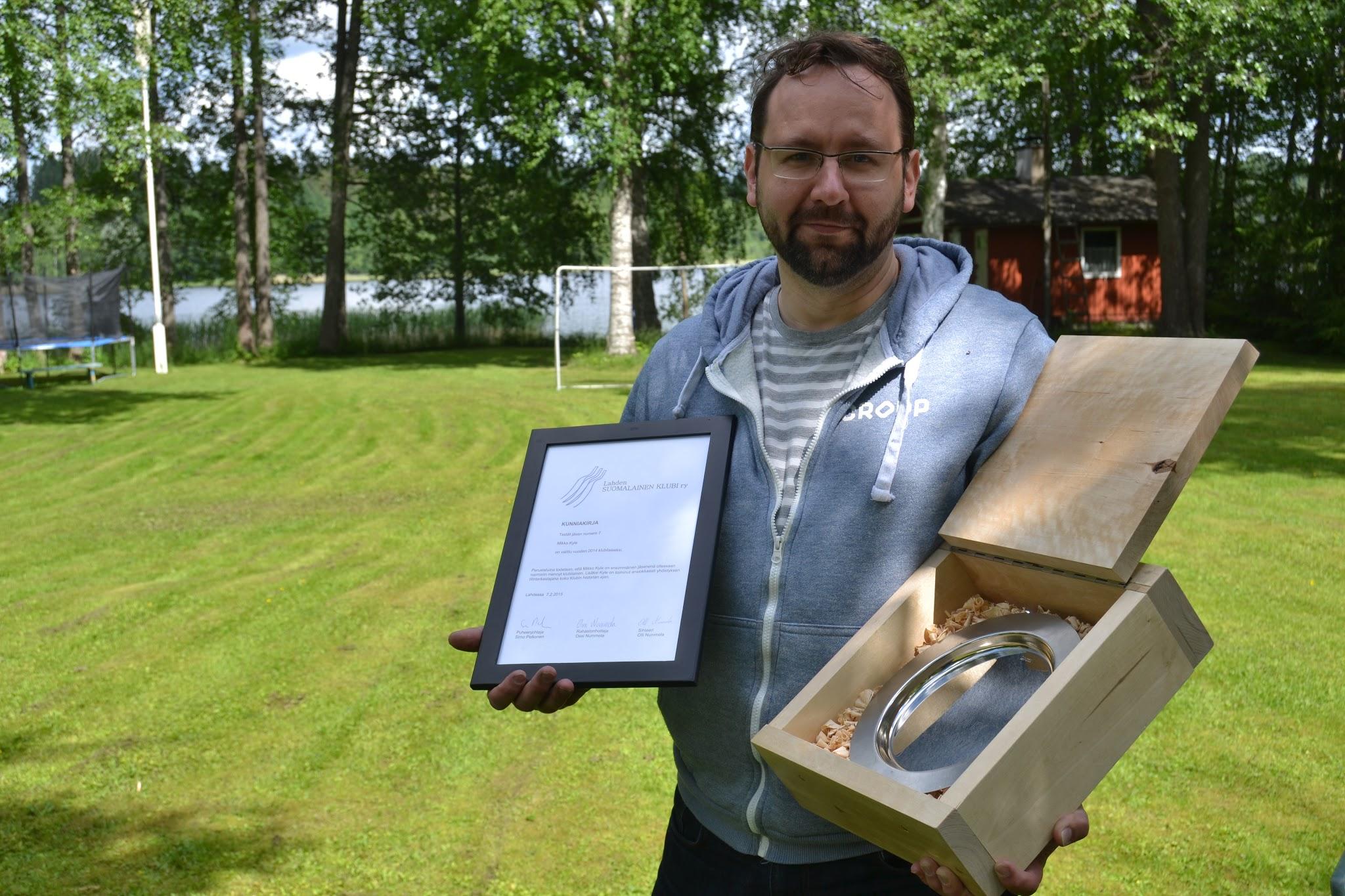 Mies pitelee kunniakirjaa, jonka mukaan Mikko Kyle on vuoden 2014 klubilainen, sekä metallilautasta puulaatikossa