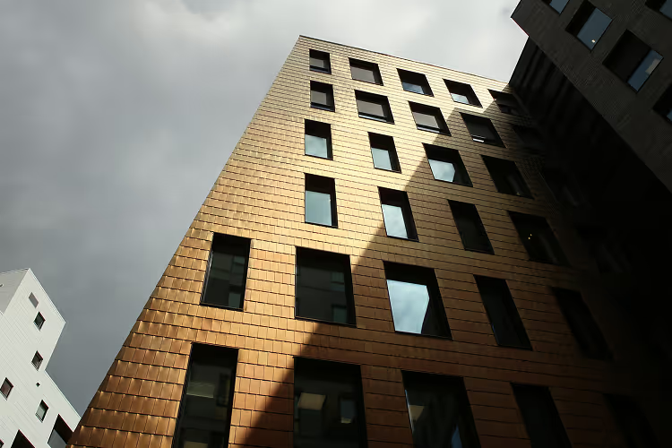 Moderne bygning med kobberfarget fasade og mørke vinduer under en overskyet himmel.