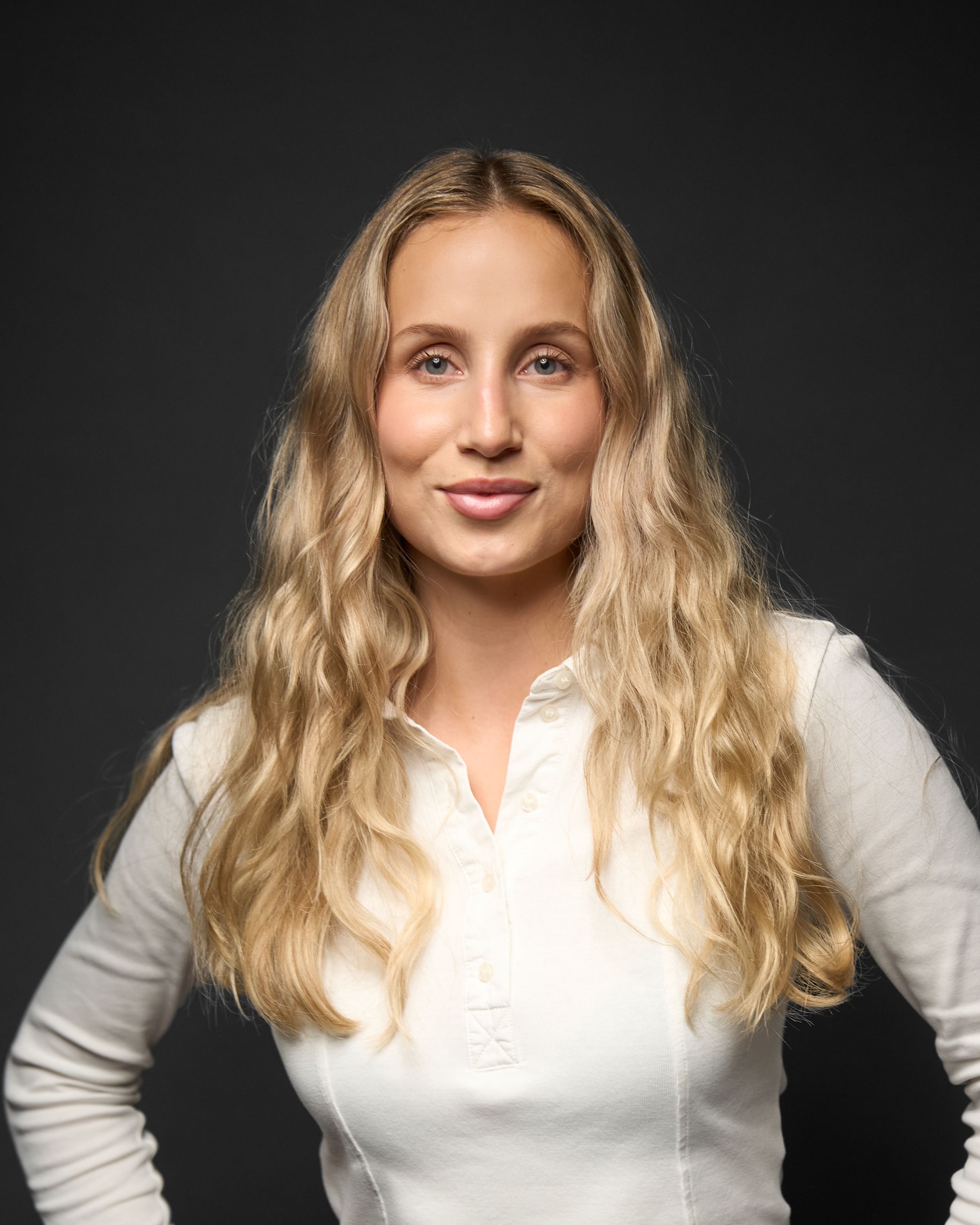 Ung kvinne med langt, bølget blondt hår iført en hvit genser med knapper står foran en mørk bakgrunn.