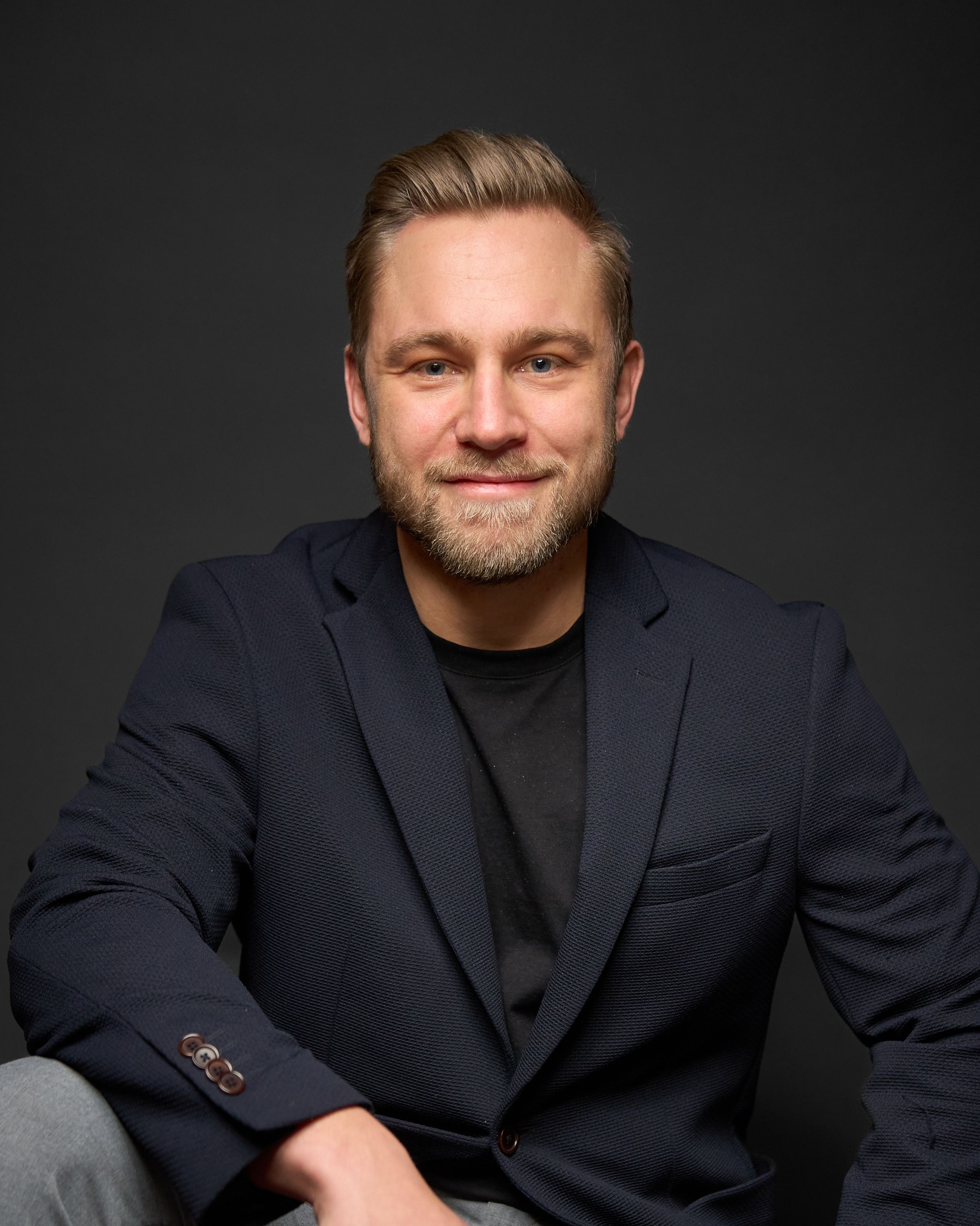 Portrett av en smilende mann med lyst hår og skjegg iført en mørk blazer og svart t-skjorte mot en mørk bakgrunn.