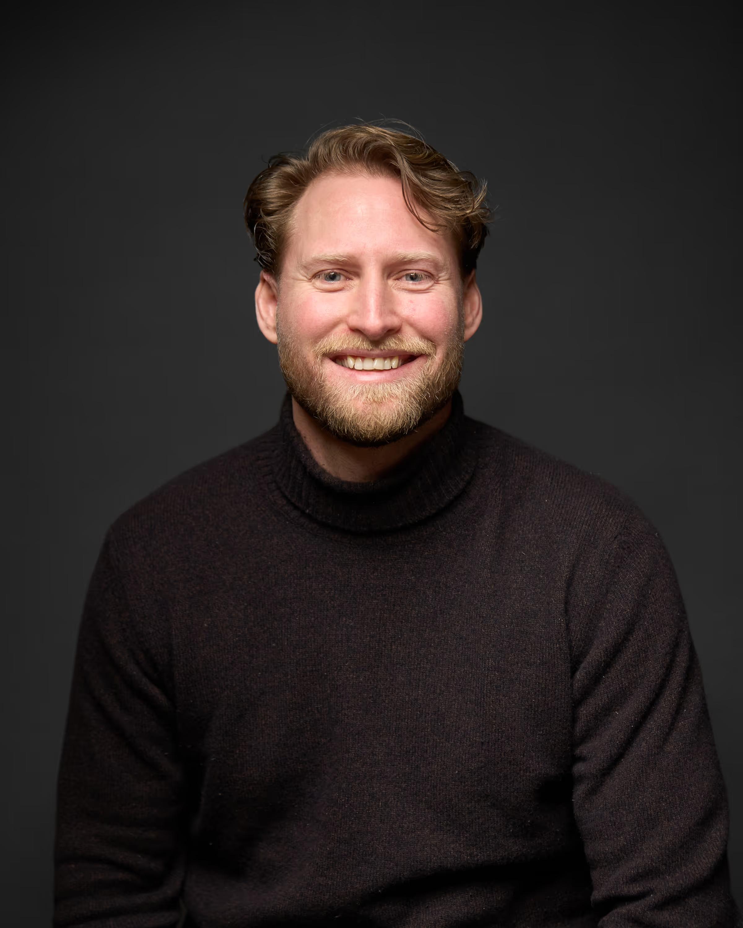 Smilende mann med lyst hår og skjegg iført mørk turtleneck-genser mot mørk bakgrunn.