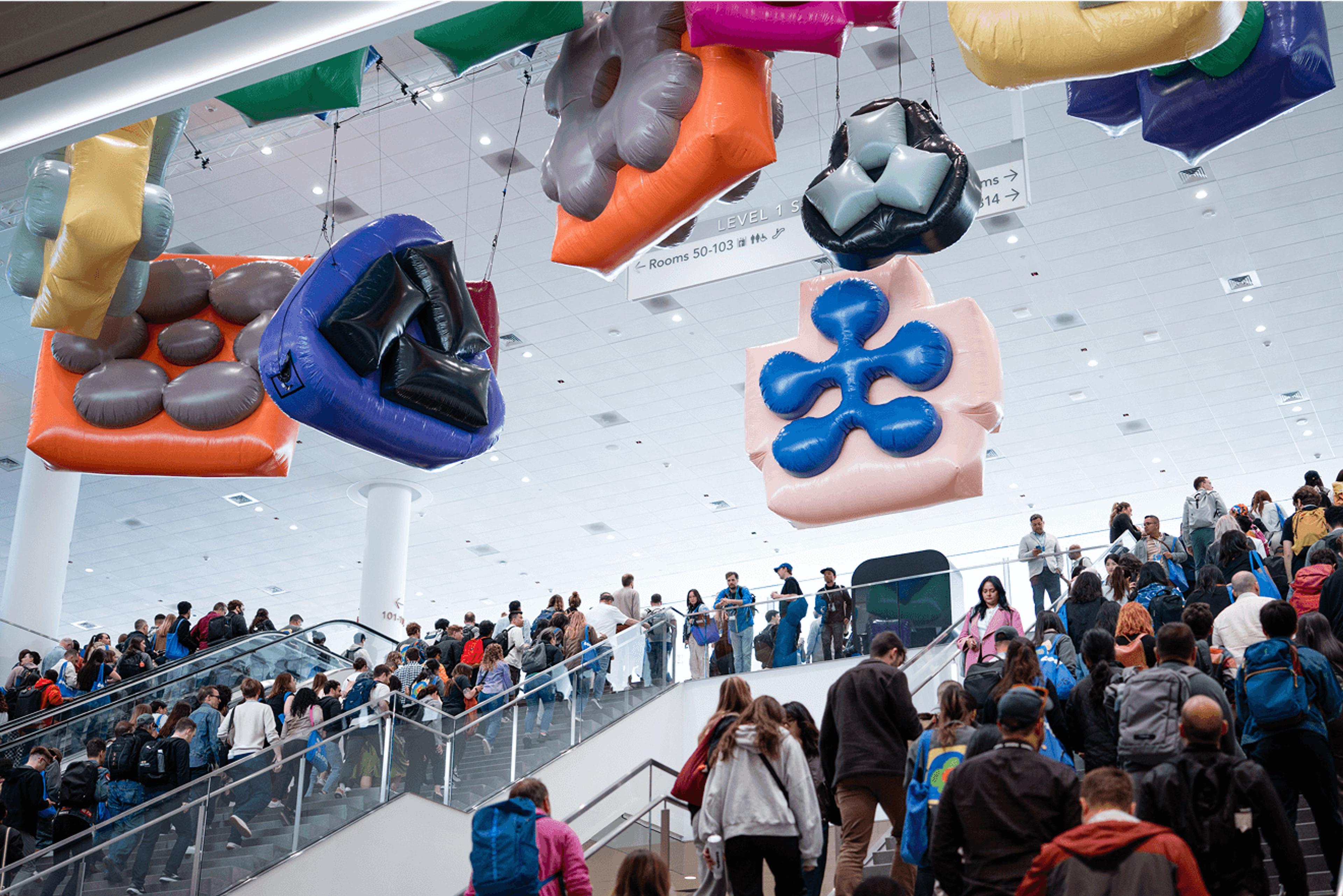Crowd moving up stairs under large inflatable Figma-shaped icons hanging from the ceiling.